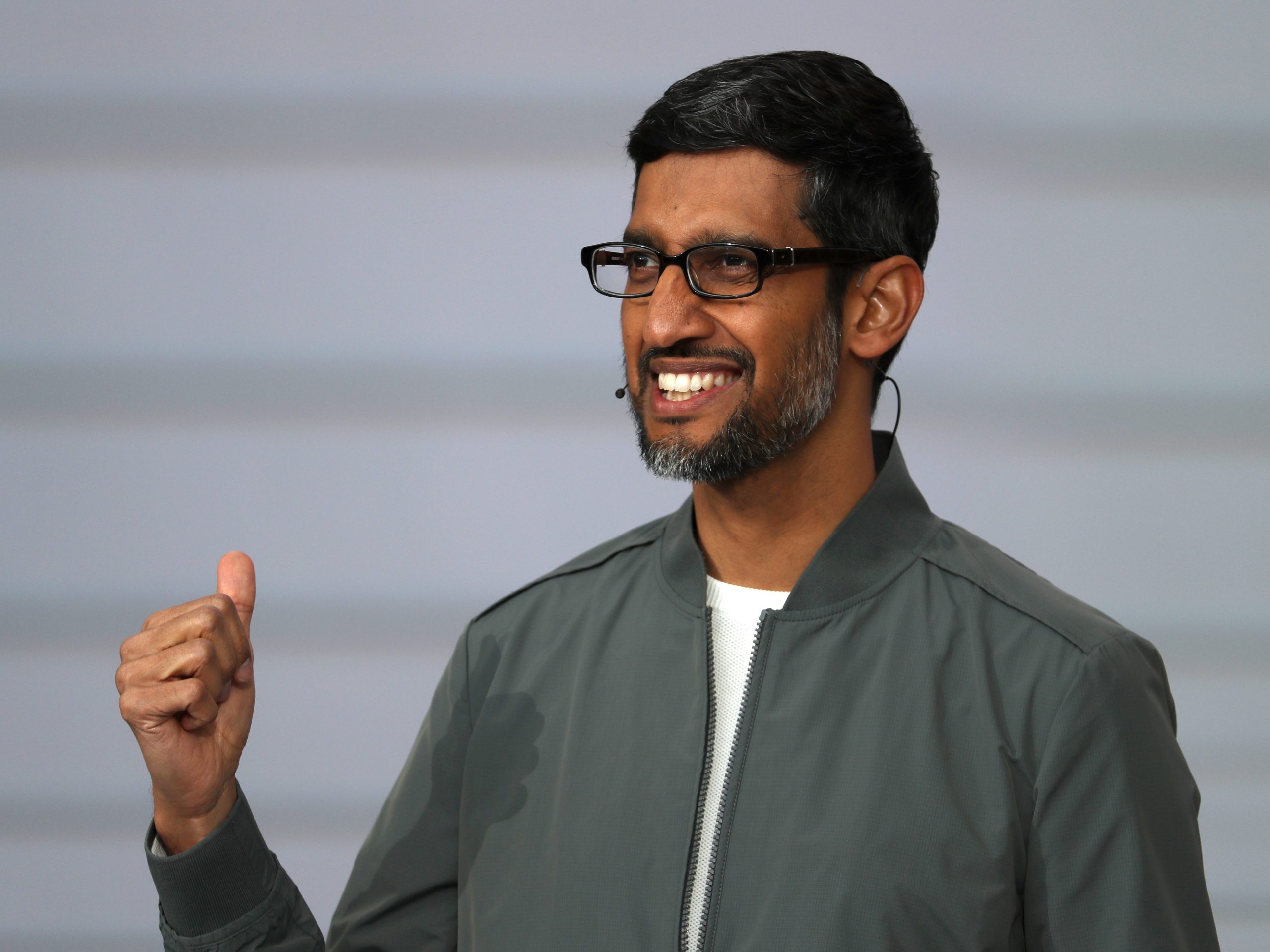 Sundar Pichai wears a grey jacket over a white t-shirt and smiles on stage.