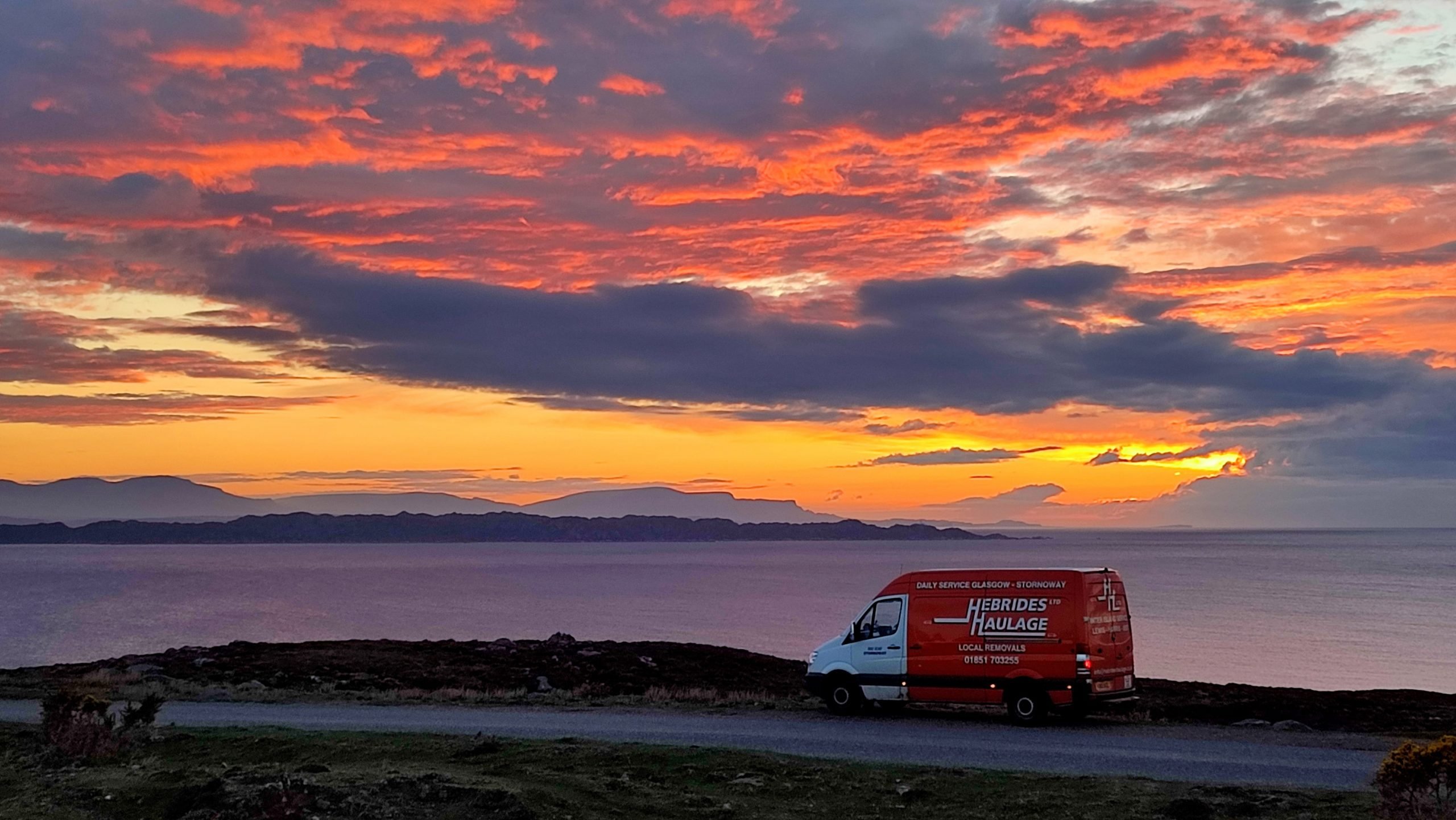 Truck driving at sunset down a coastal road in Scottish Highlands.
