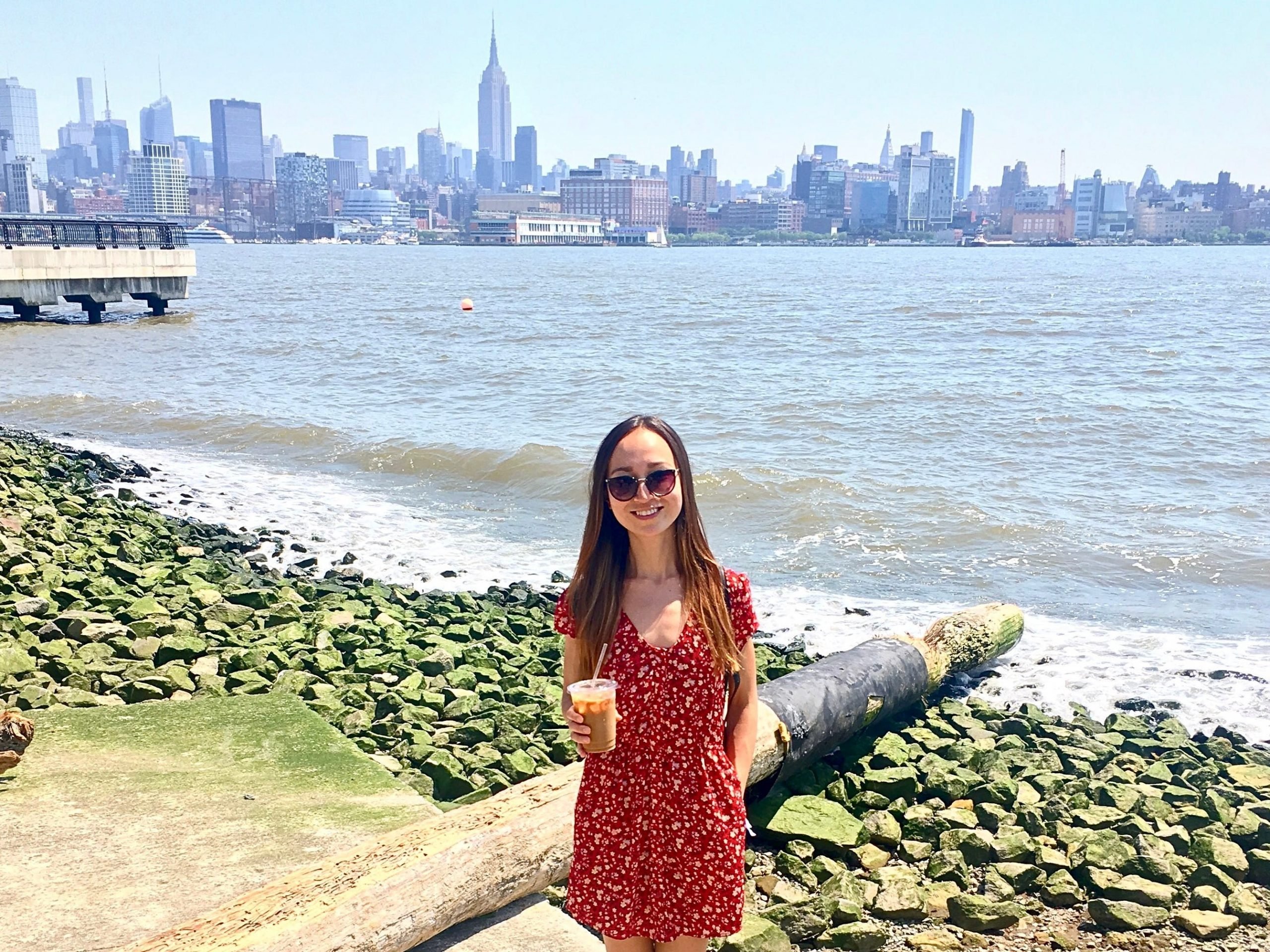 The writer standing in front of the water holding a drink with the new york skyline in the background