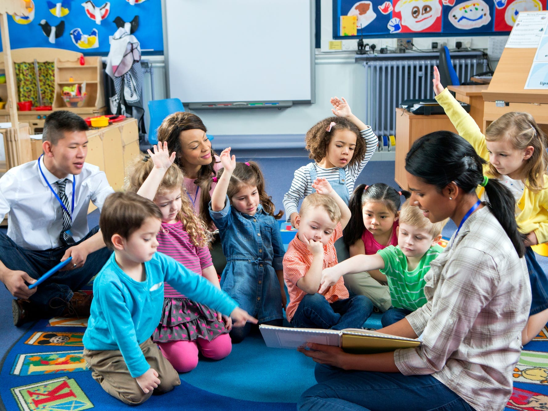 A teacher is reading to students. The teacher, children, and two other teachers are sitting on the floor and some children have their hands up to ask a question.