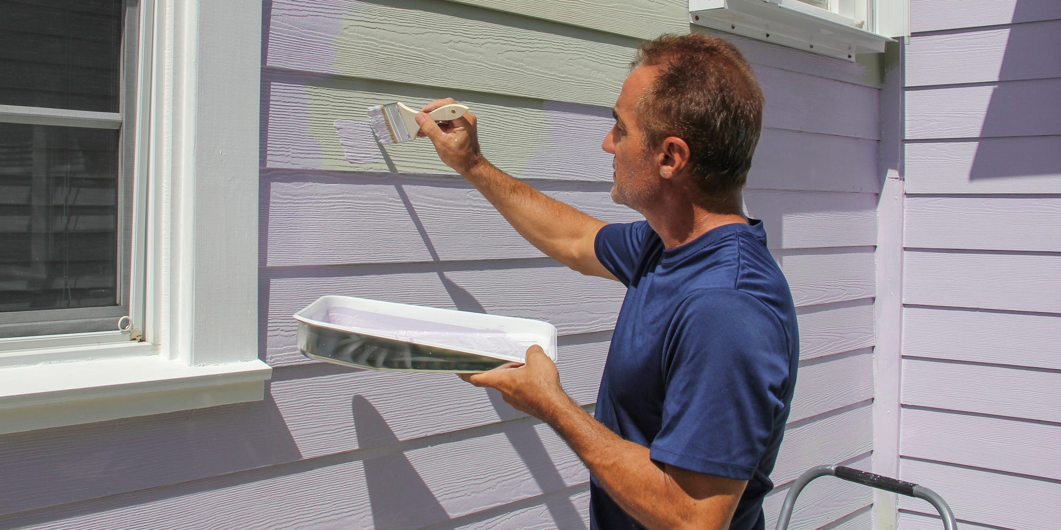 A man handing a tray of paint and a paintbrush as he paints the siding on a house from gray to purple