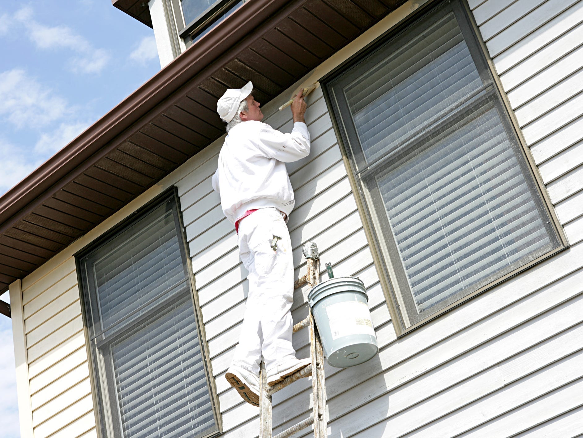 A painter on a ladder painting siding with a brush