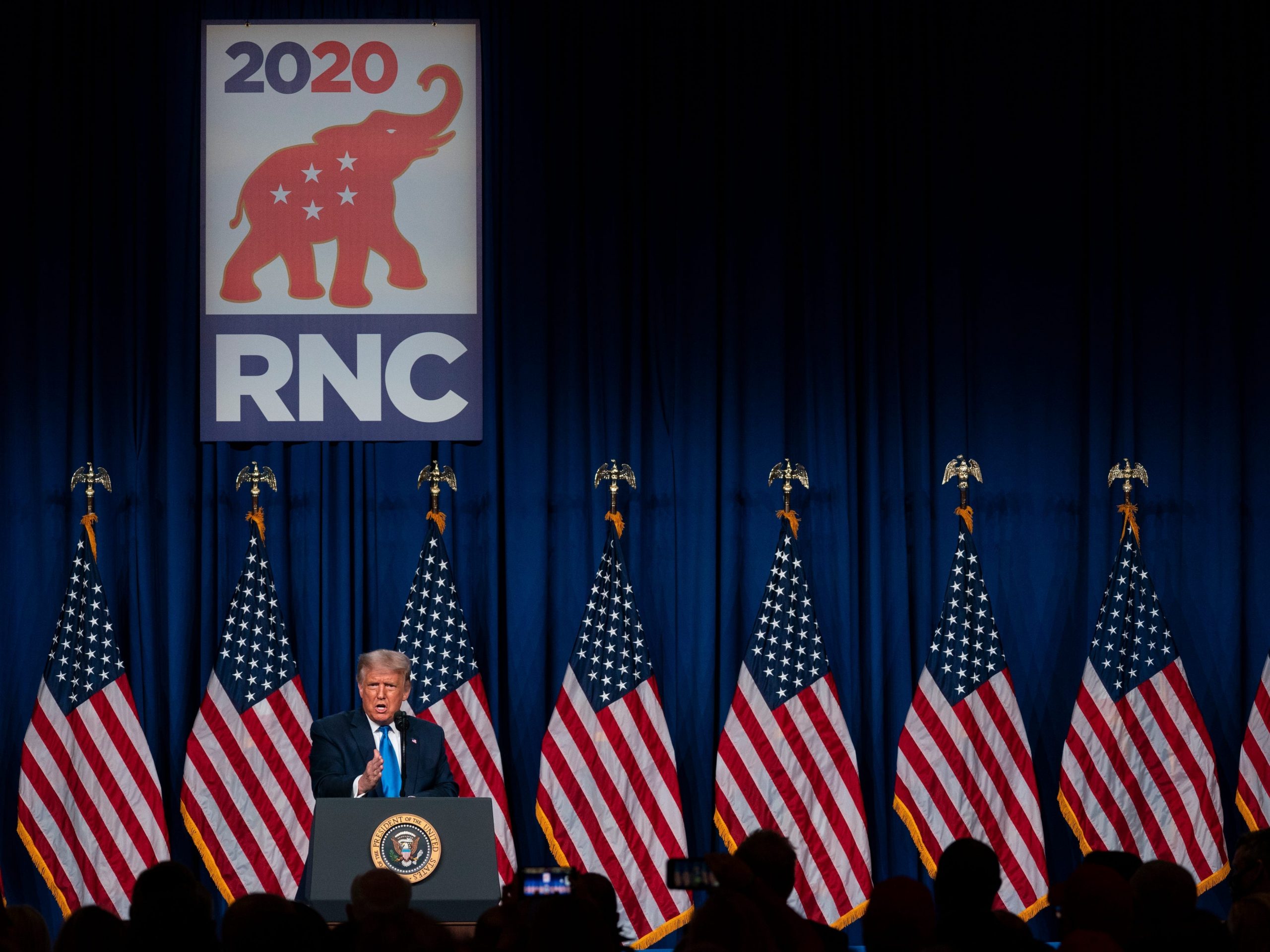 Donald Trump speaks in front of an RNC sign and many American flags.