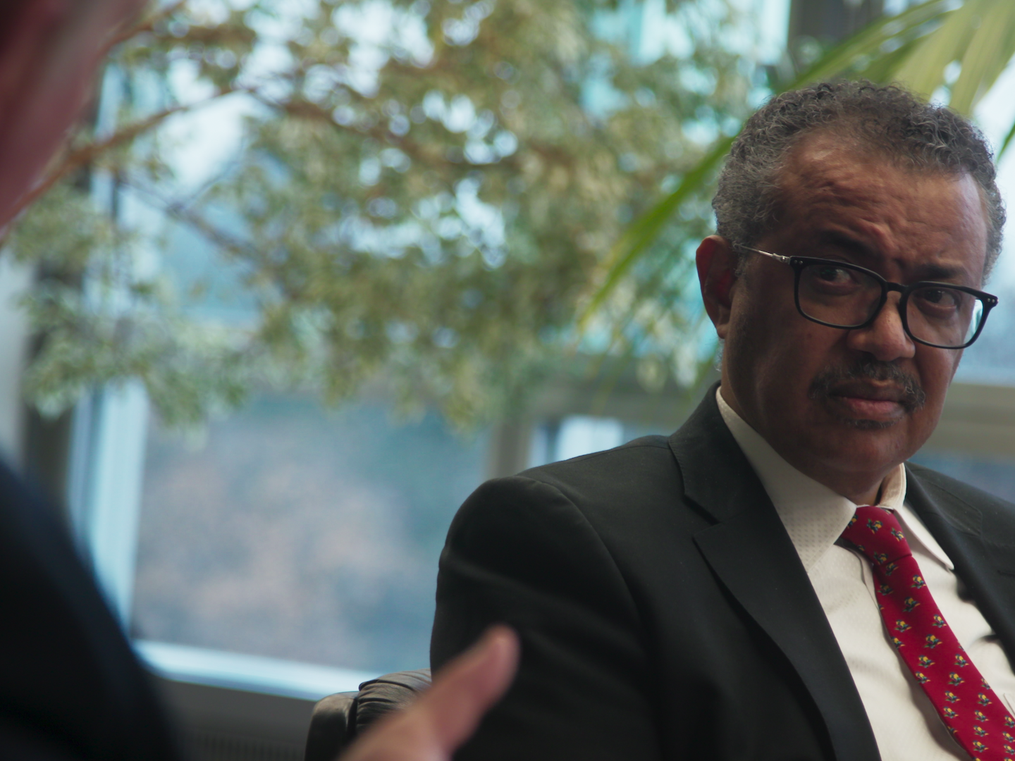 Tedros sitting down in a suit and tie, listening to his colleague Mike Ryan speak.