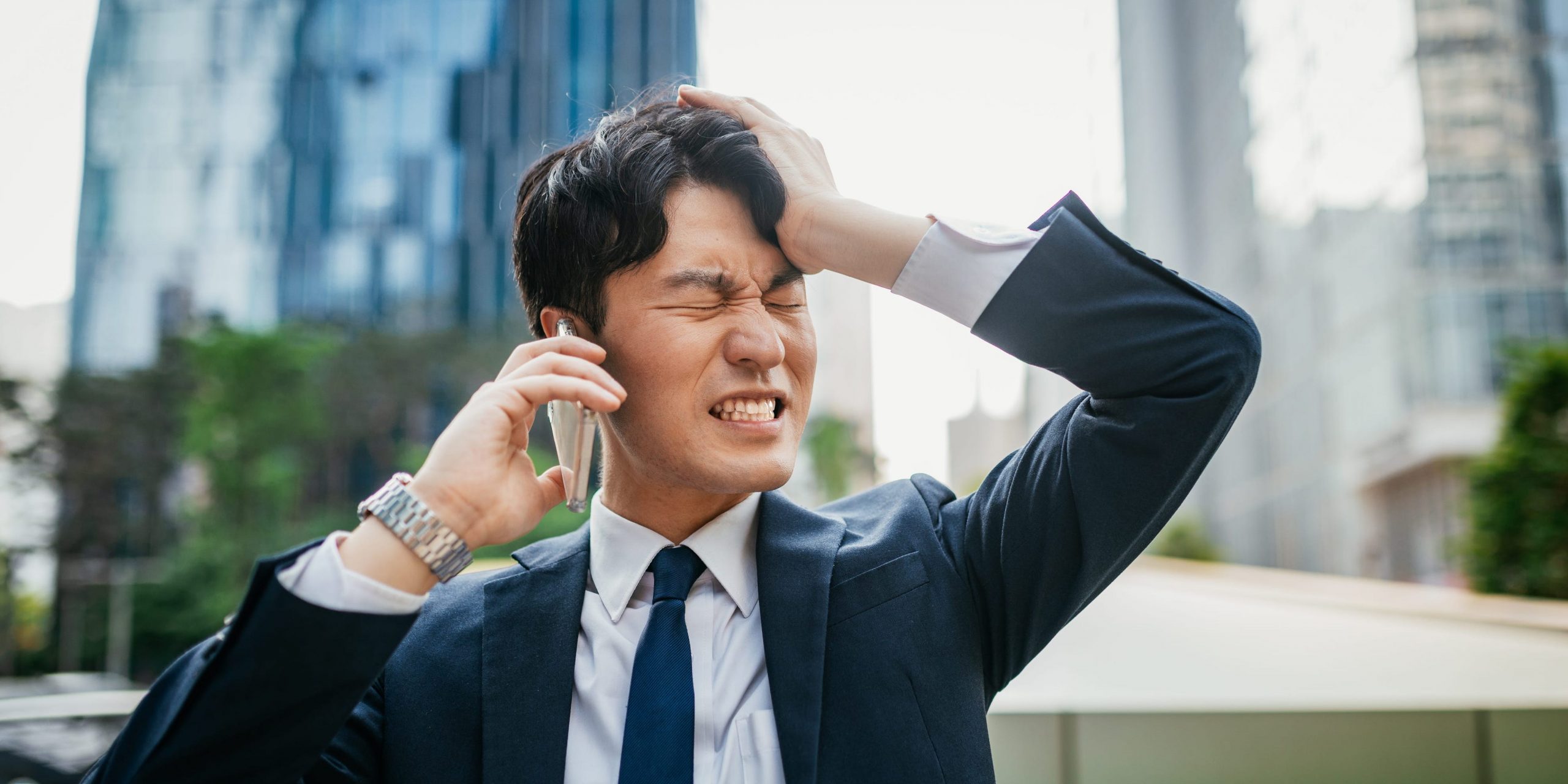 Business man in a suit, gripping his head while on the phone outside.