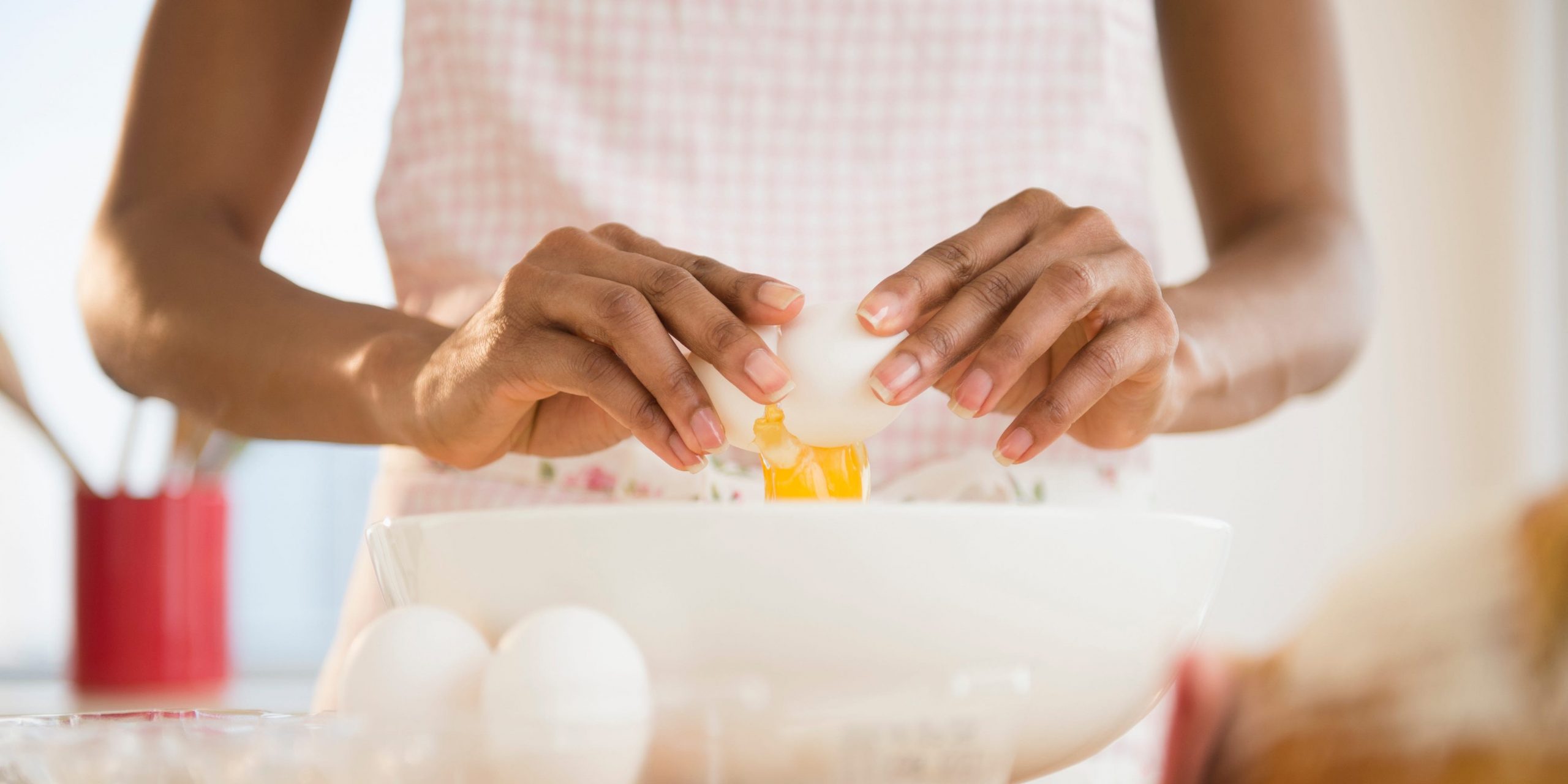 A person wearing a pale pink checkered apron cracks a raw egg into a white bowl.