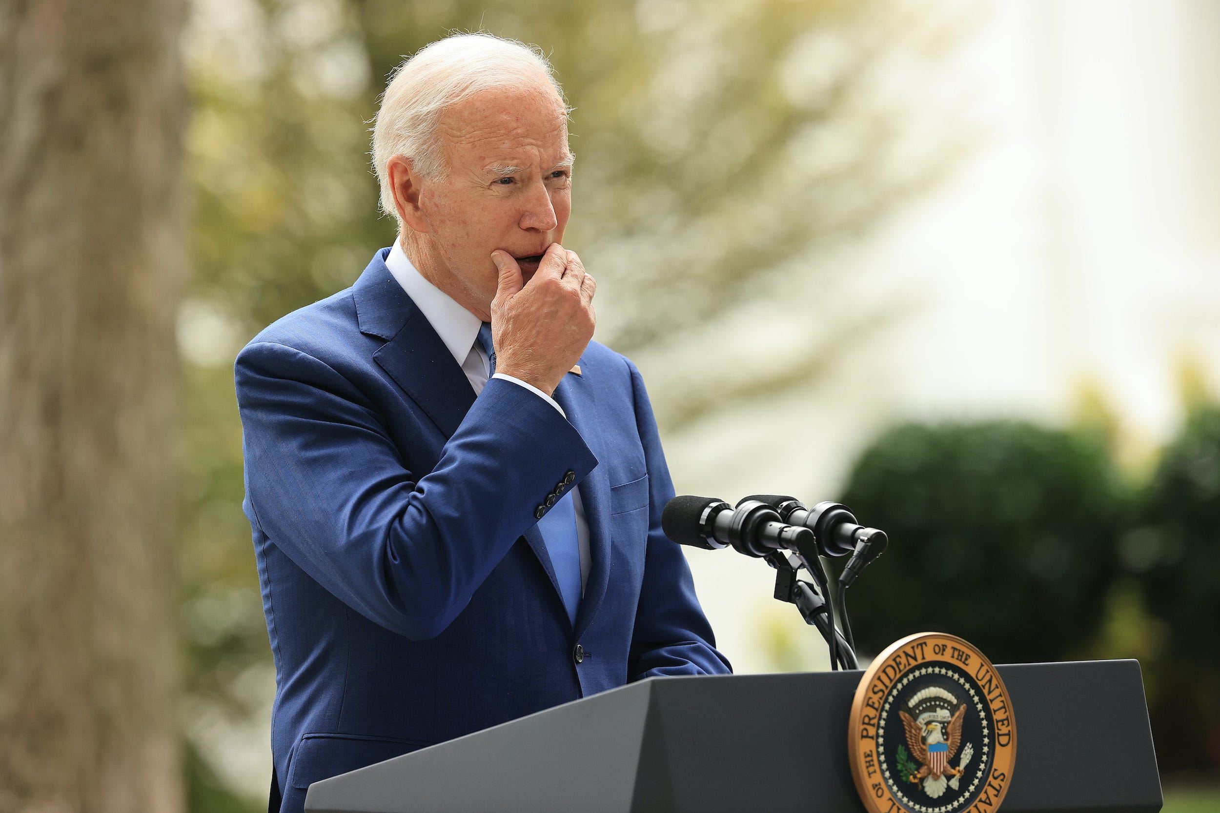 President Joe Biden stands outside behind a podium with the presidential seal, grabbing his chin and slightly covering his mouth in a pensive look.
