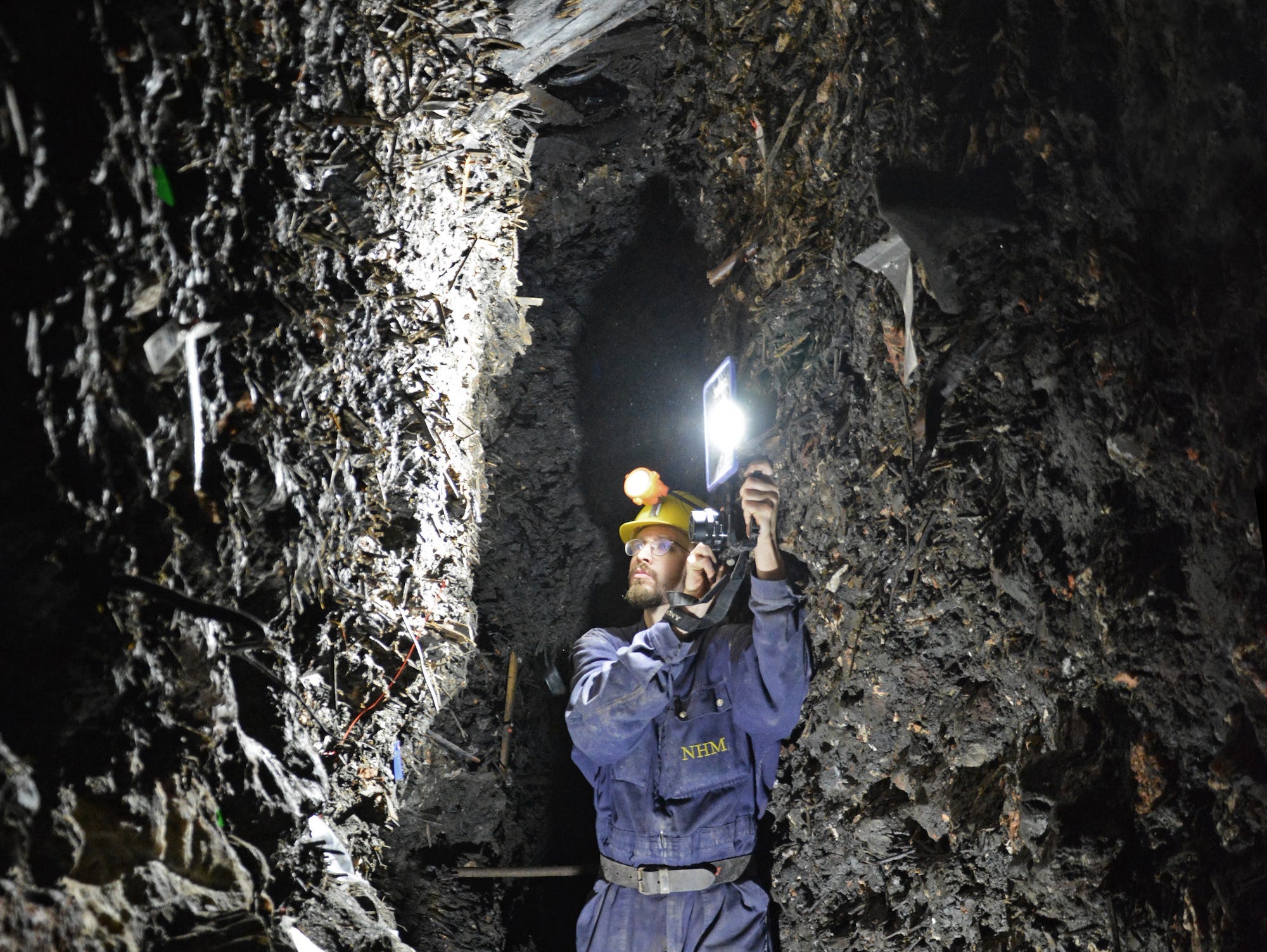 A man wearing a yellow helmet and blue jumpsuit holds up a light in a narrow rock passage in the Hallstatt mine.