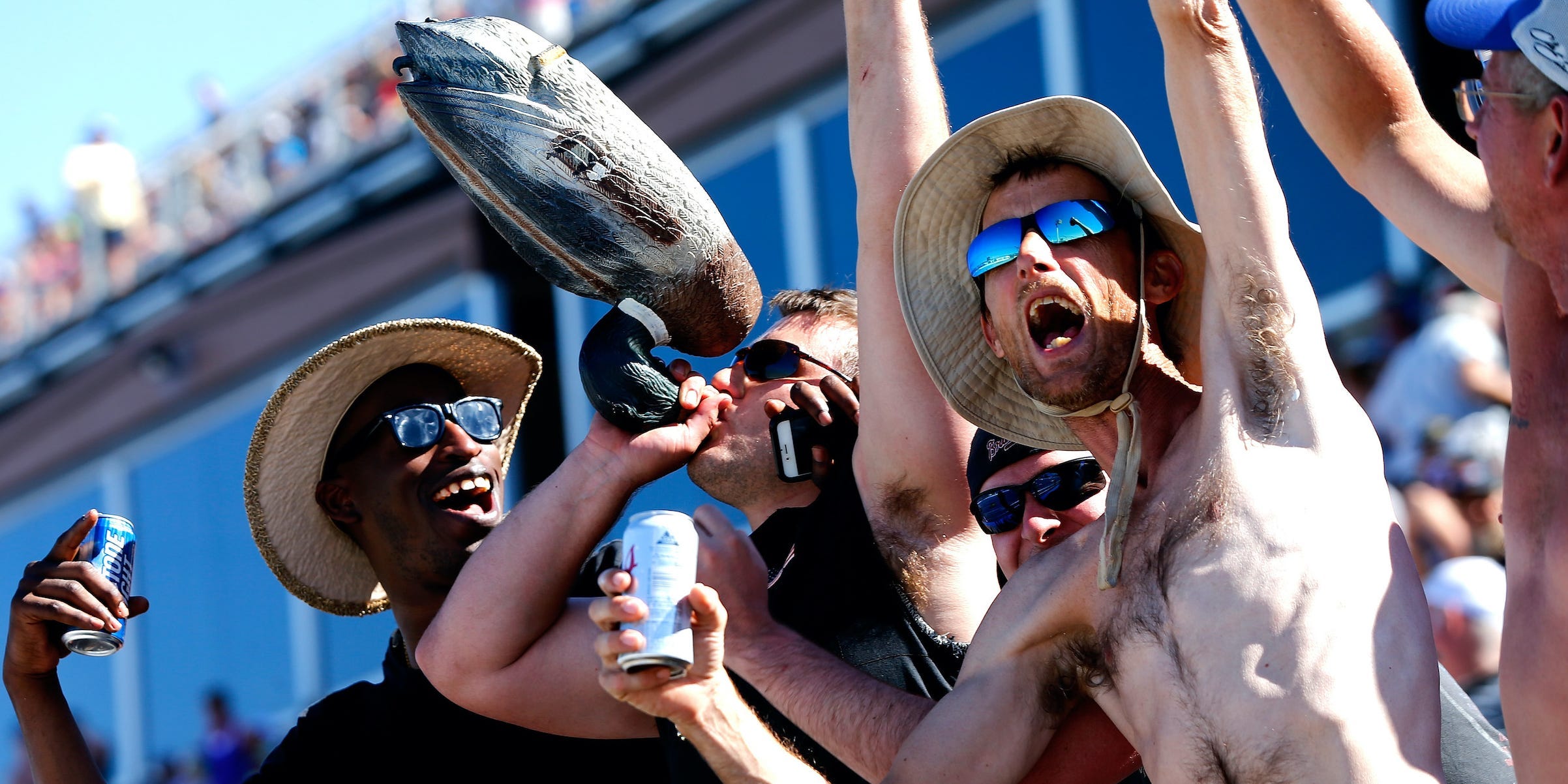 NASCAR fans cheer with their arms in the air. One man, shirtless, holds a beer, while another appears to drink from the bill of an upside down duck.
