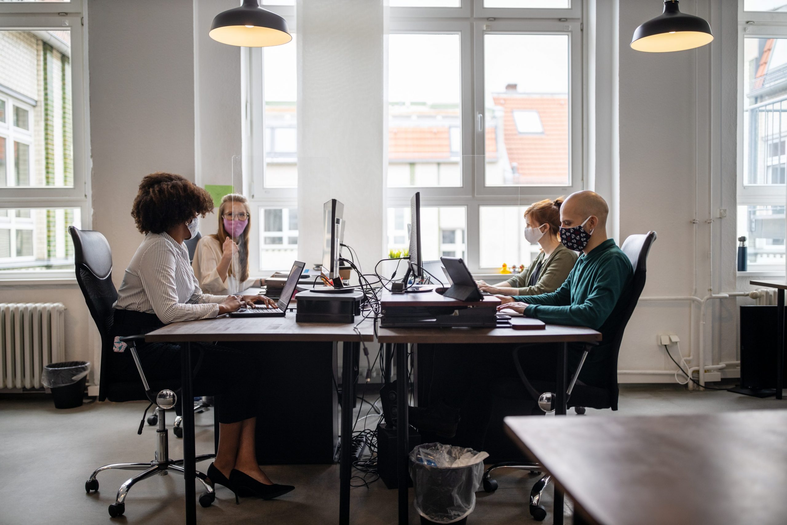 Four coworkers wearing masks sit in an office together, talking and working on laptops