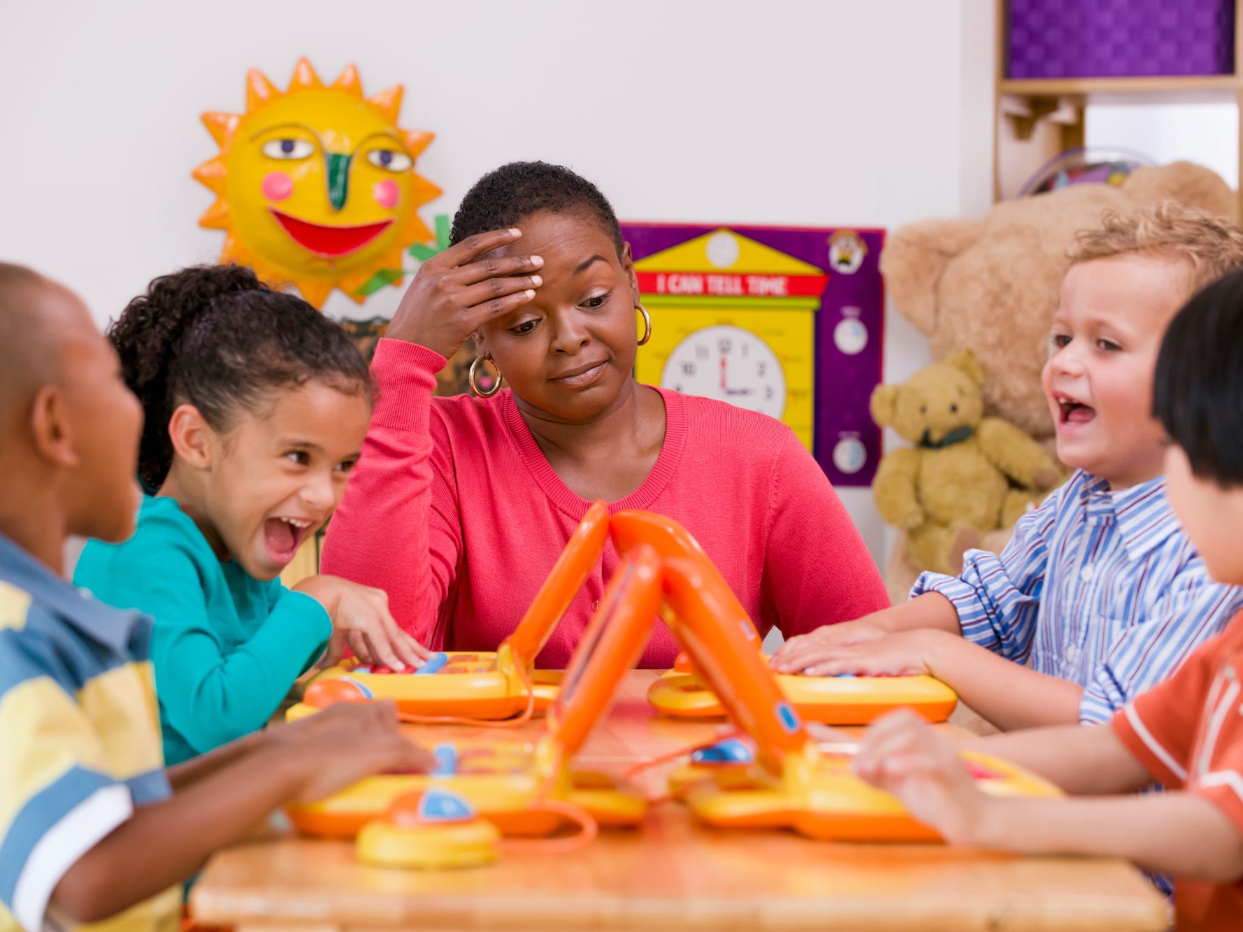 Four children playing with toy laptops in a class. A female teacher is sitting with them at the table