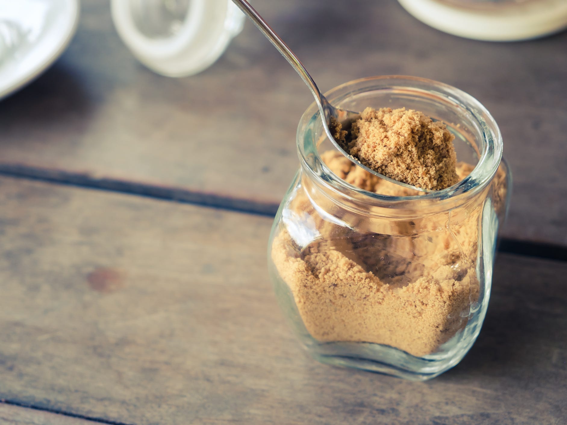 A glass jar of brown sugar with a spoon in it sitting on a wooden table