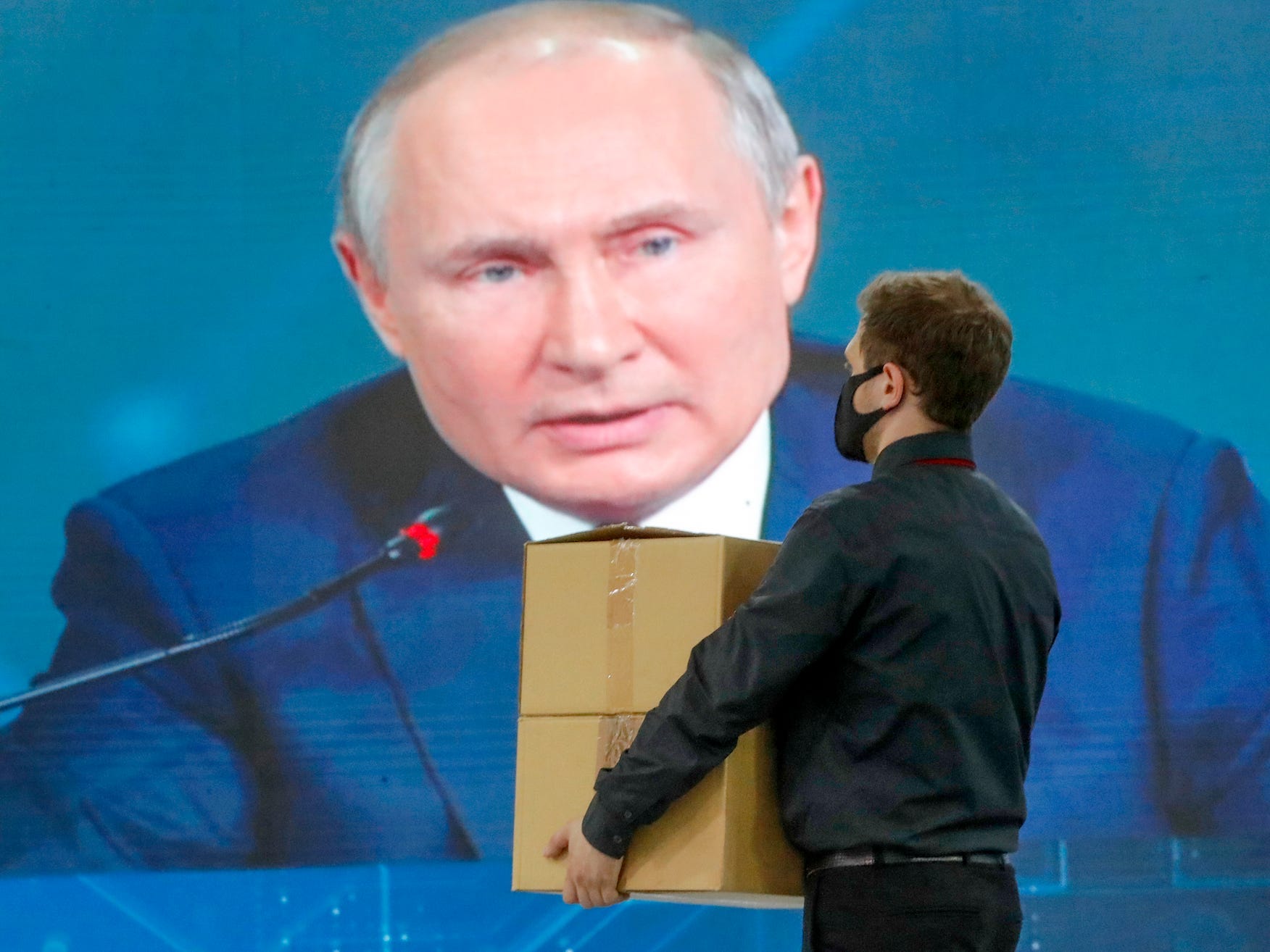 aman carrying boxes and wearing a mask walks in front of a giant screen showing President Vladimir Putin.