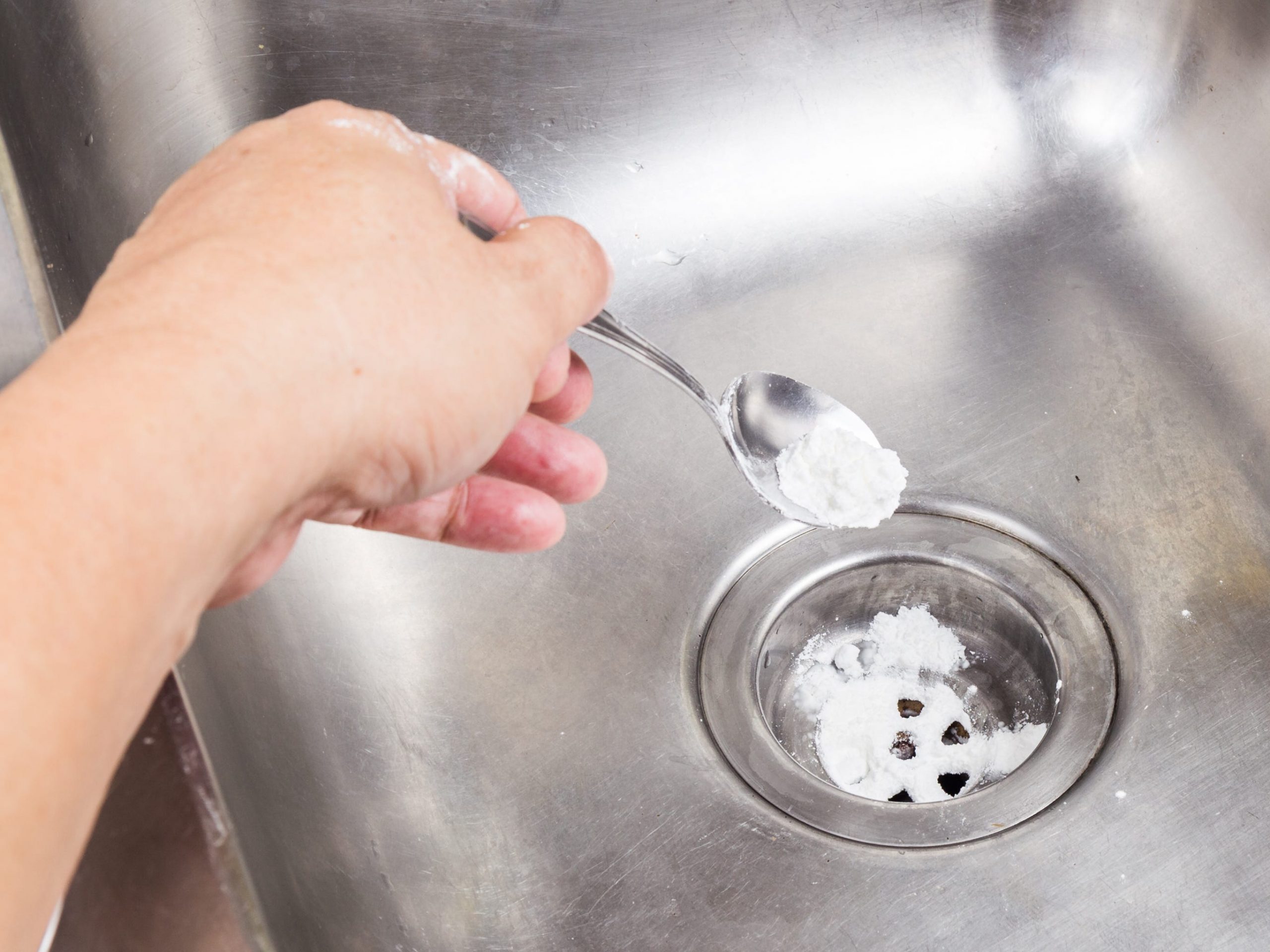 A person's hand using a spoon to pour baking soda into a sink drain