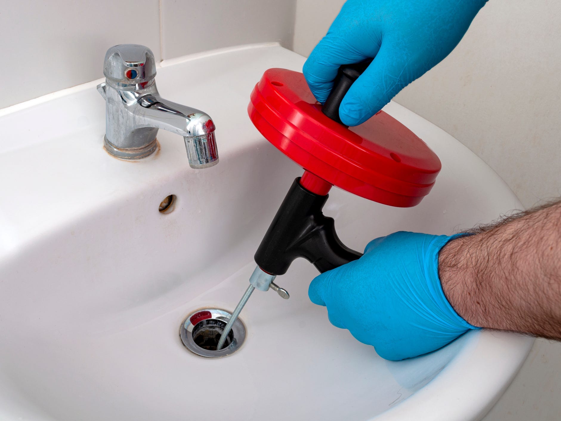 A pair of gloved hands holding onto a drain snake that's been inserted into a sink drain