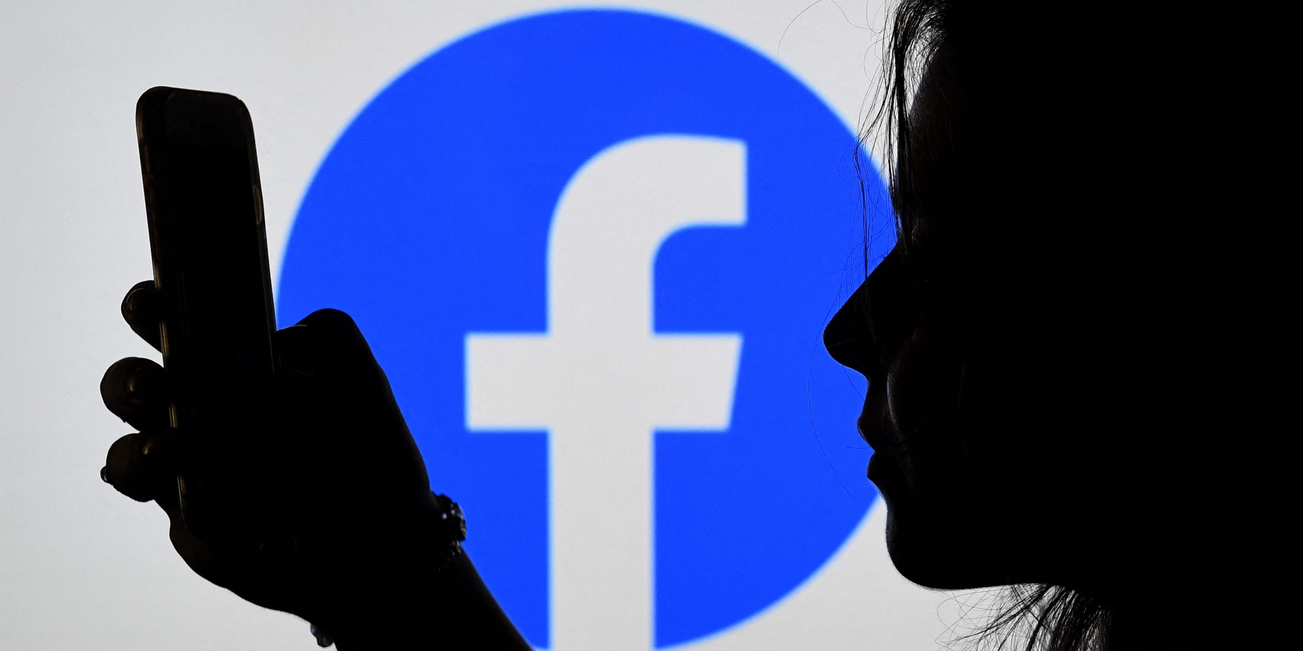 A person looks at. a smart phone with a Facebook logo displayed in the background