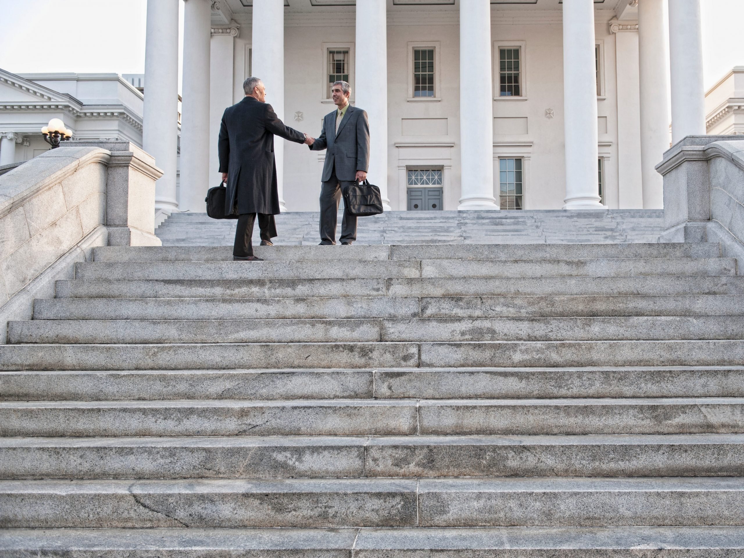 Stock photo of two men in business suits shaking hands on the steps to a courthouse.