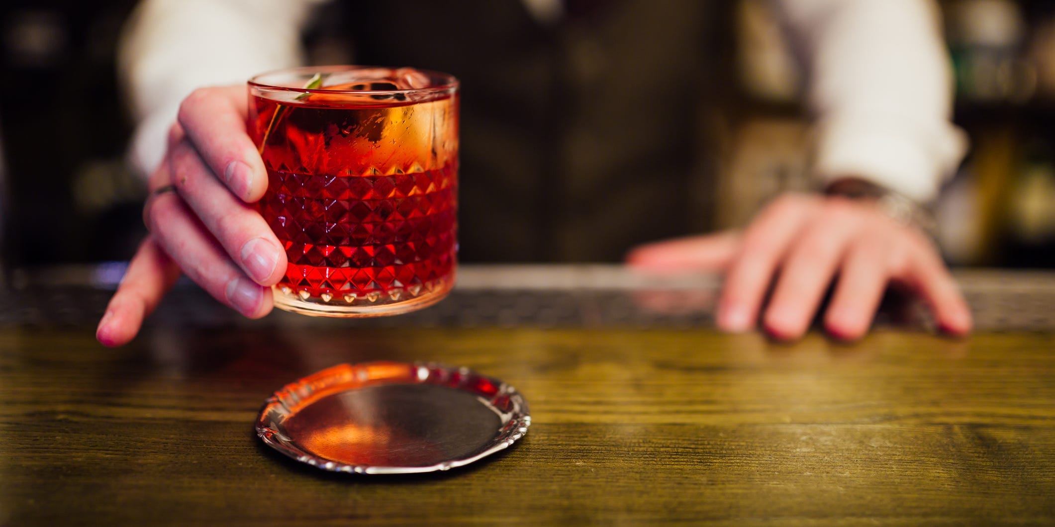 A bartender placing a negroni onto a silver coaster on a wooden bar