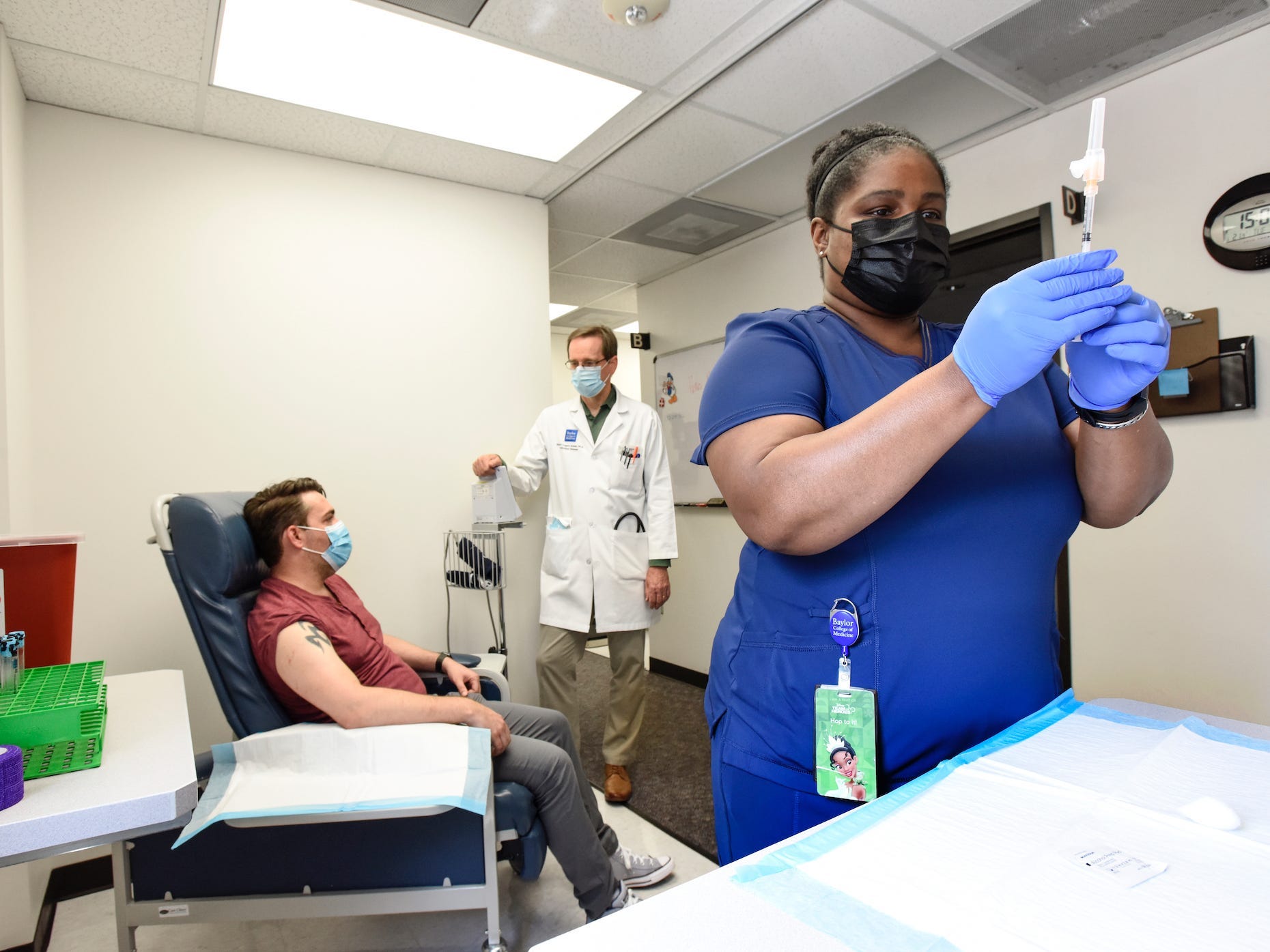 woman in scrubs prepares covid-19 vaccine, with patient and doctor chatting in background