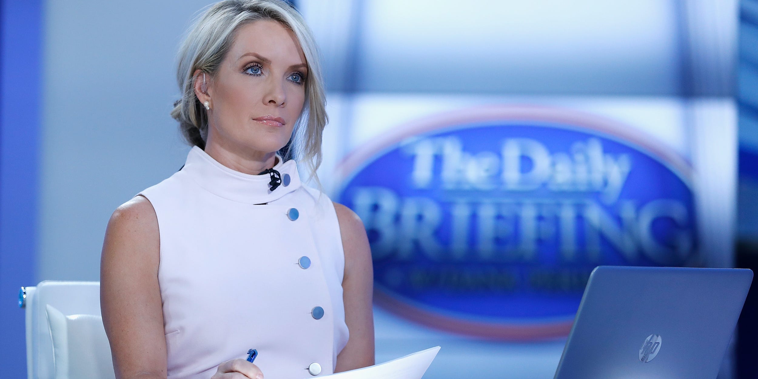 Fox News host Dana Perino sits on set in front of a laptop with notes in her left hand and a pen in her right.