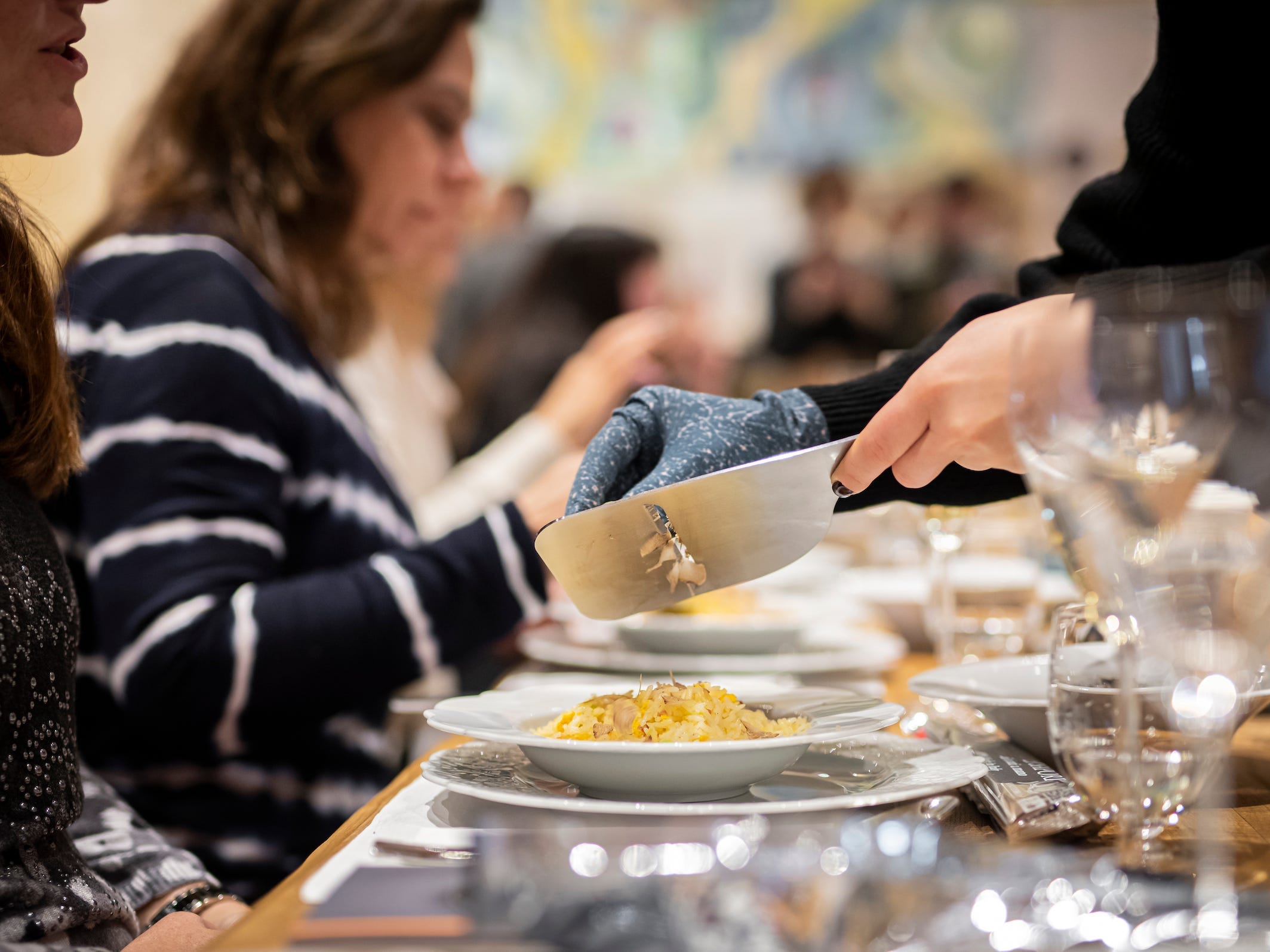 Waiter shaves white truffle onto pasta for two women sitting at table among group of diners