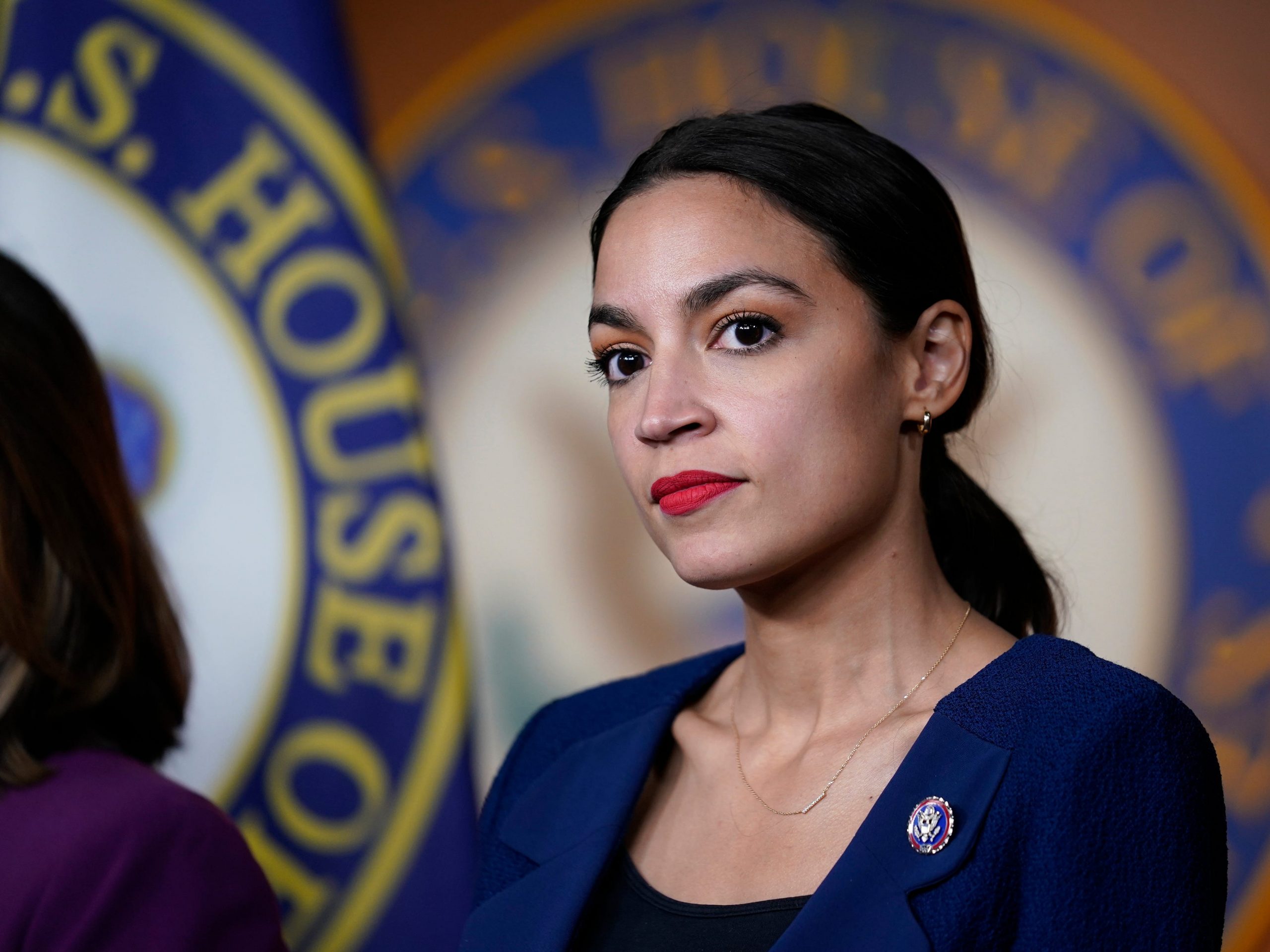 New York Congresswoman Alexandria Ocasio-Cortez in a blue blazer in front of a House of Representatives seal