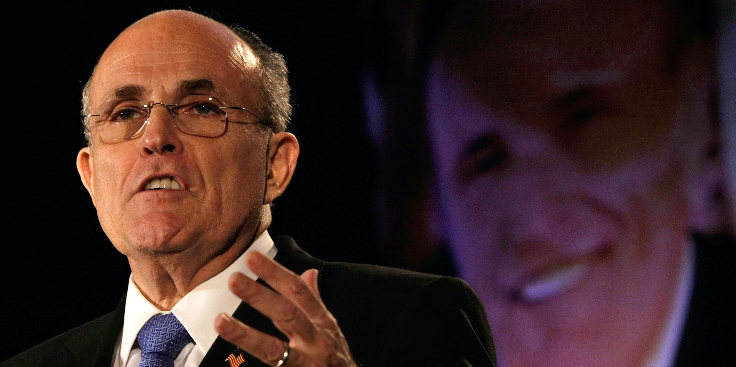 Rudy Giuliani speaks in front of a projected photo of his face.