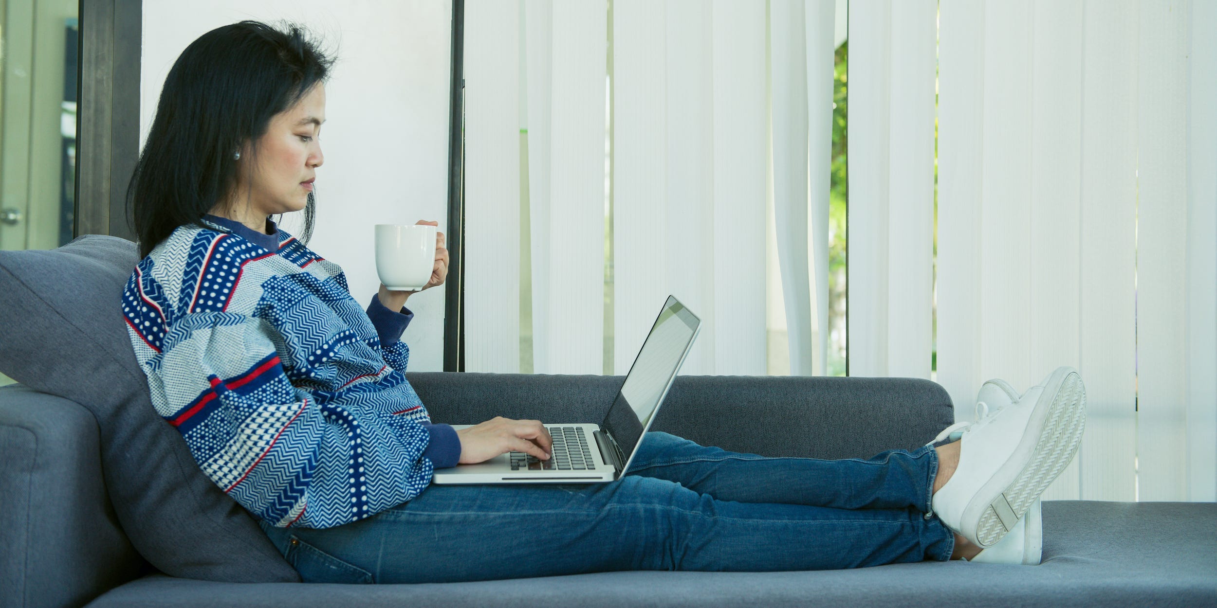 woman using laptop computer on couch at home with coffee in hand