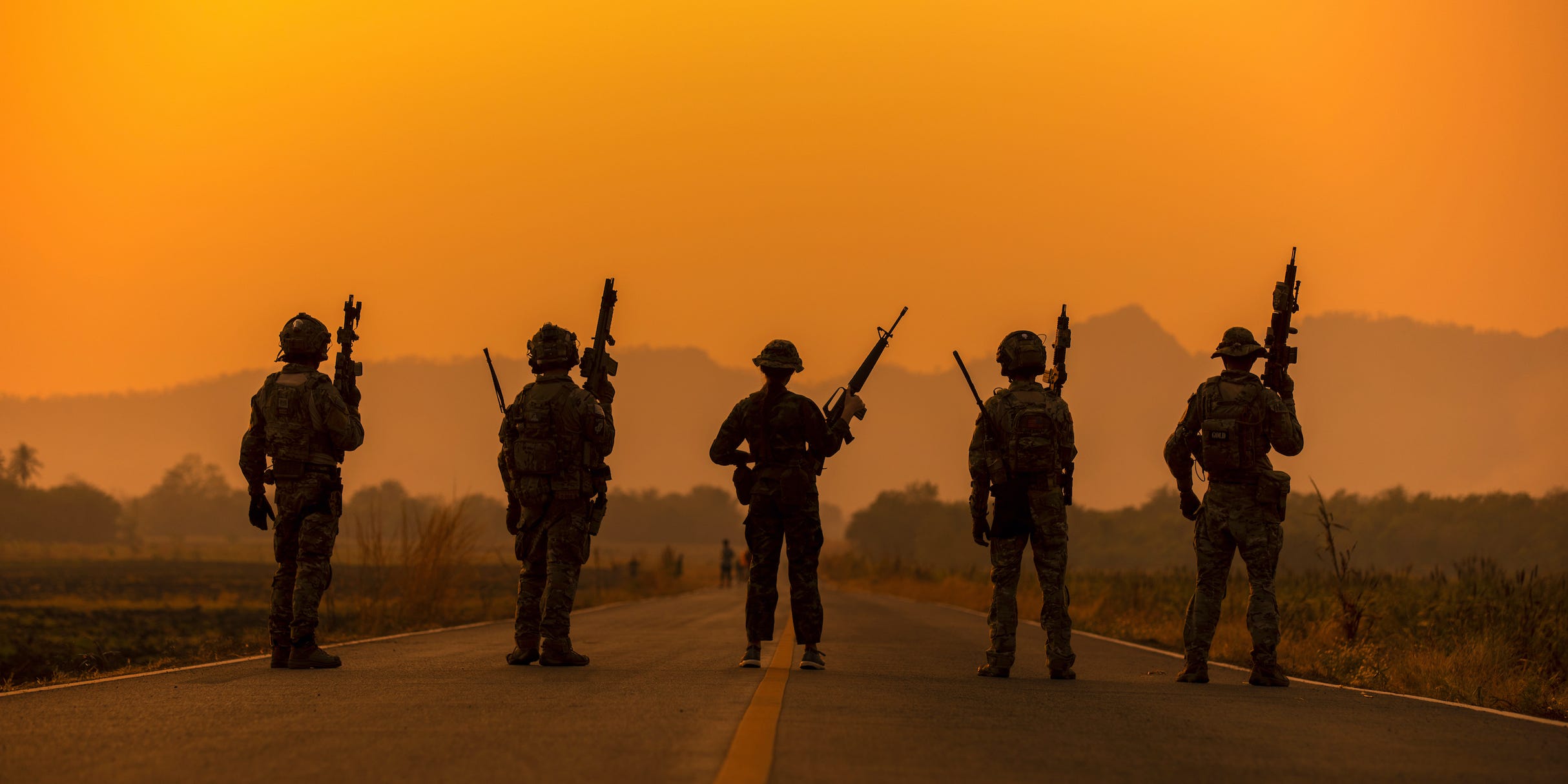 Five soldiers stand shoulder to shoulder in the middle of a road, facing a hazy sunset over mountains in the distance.