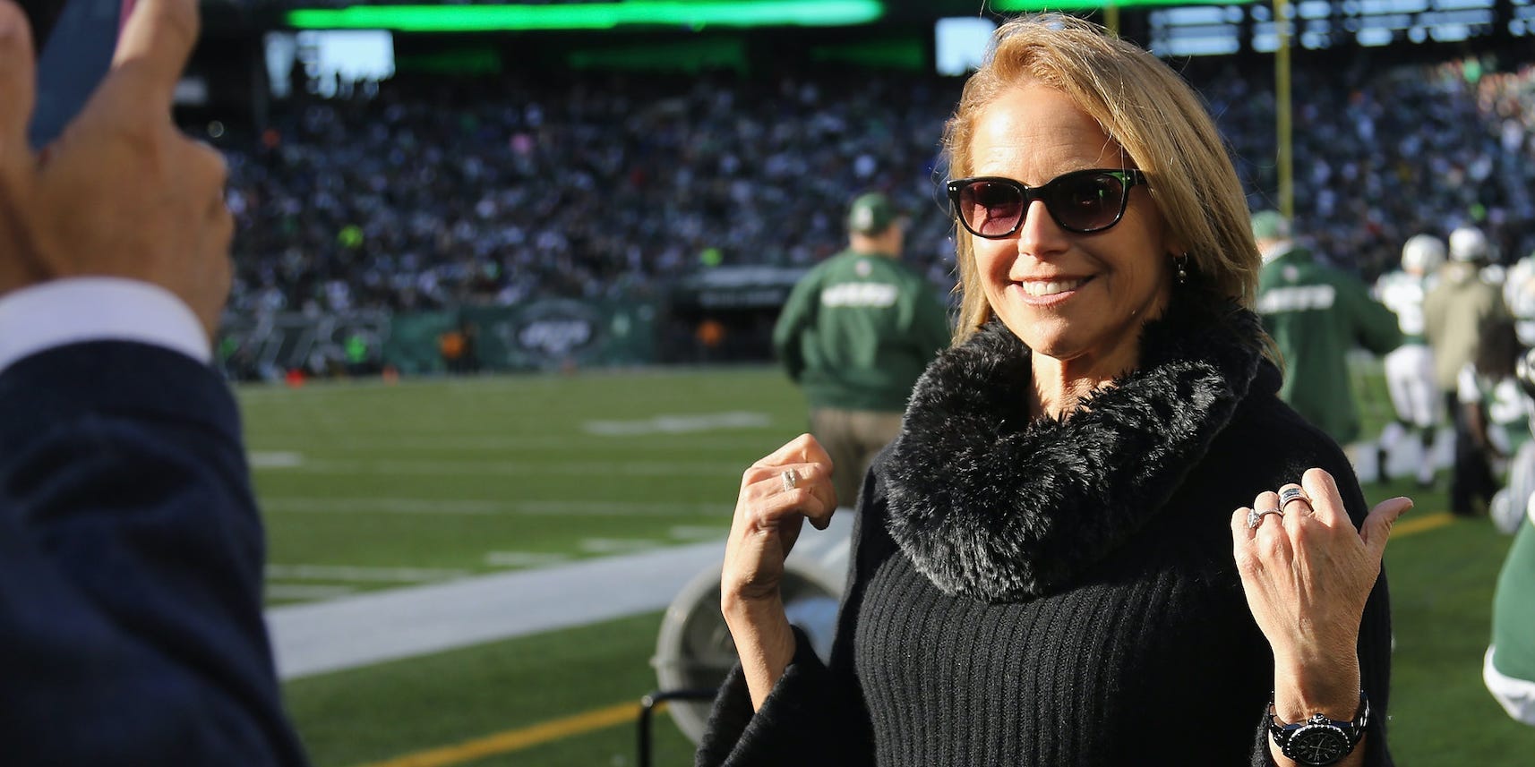 Katie Couric stands off the sidelines at MetLife Stadium for a New York Jets game, wearing a black sweater and sunglasses while pointing her thumbs behind her at the players in posing for a picture.