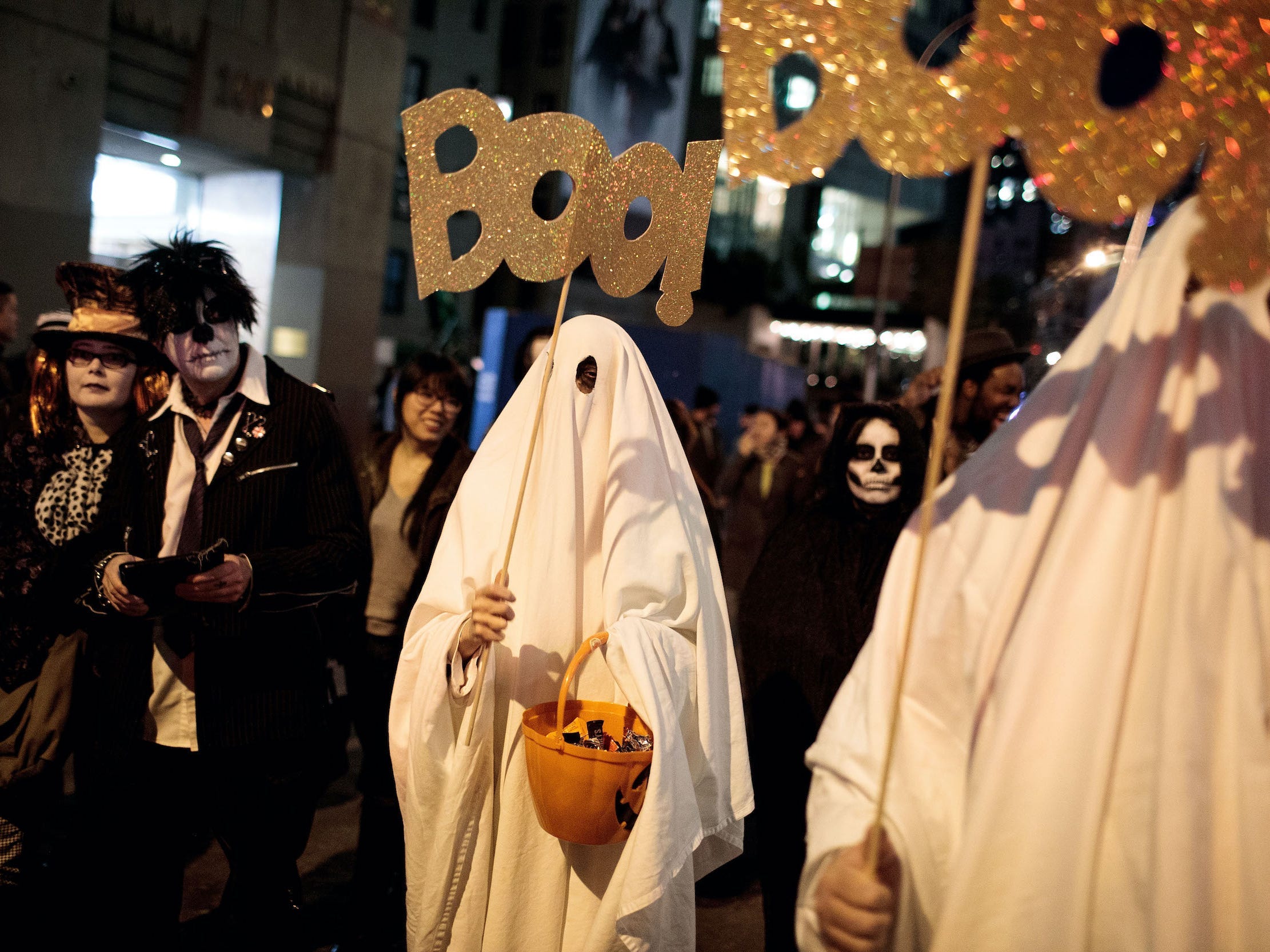 Person dressed as ghost holds pumpkin and sign that says boo during Halloween parade