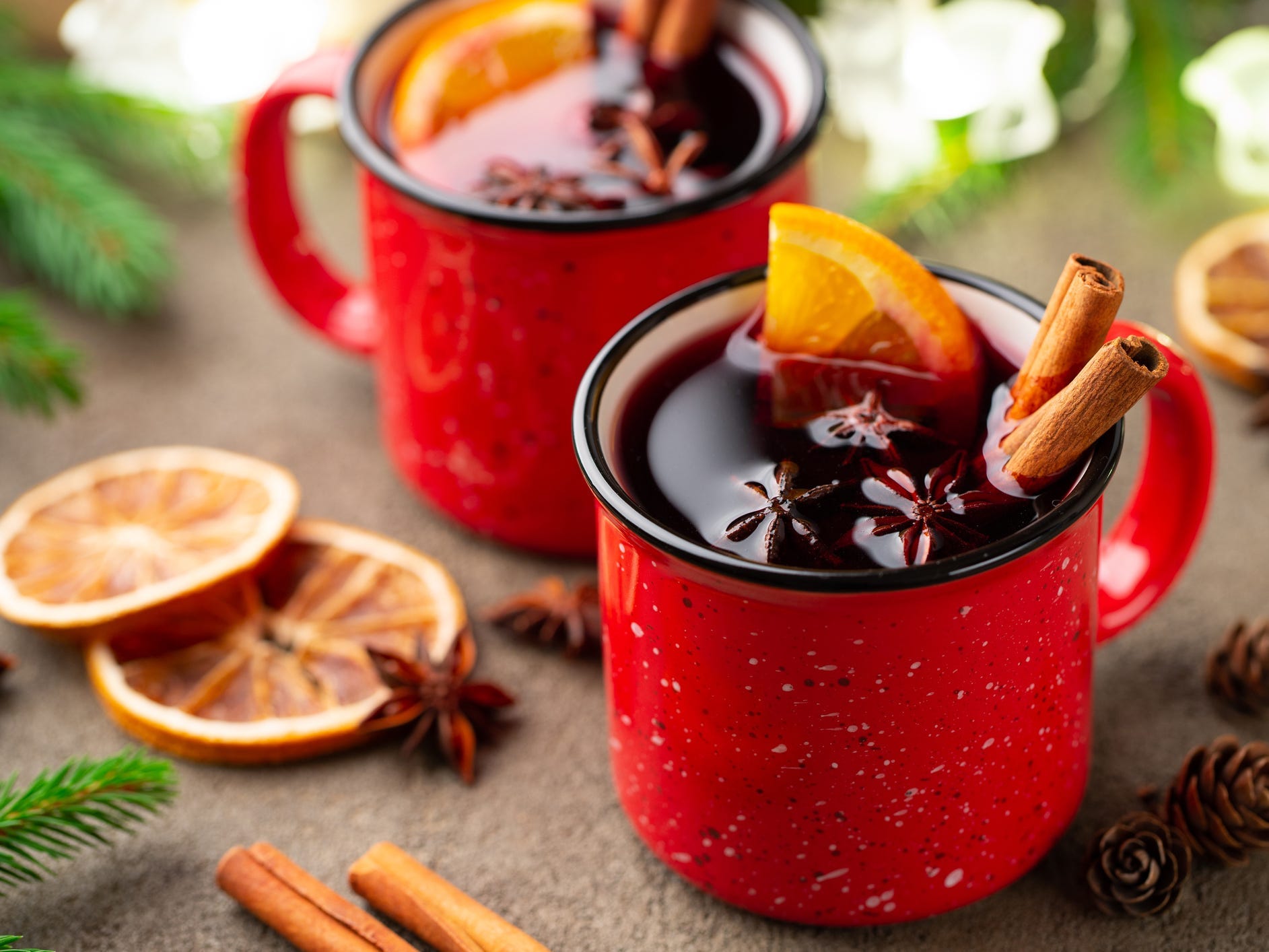 Two red mugs of mulled wine garnished with cinnamon sticks, star anise, and an orange slice