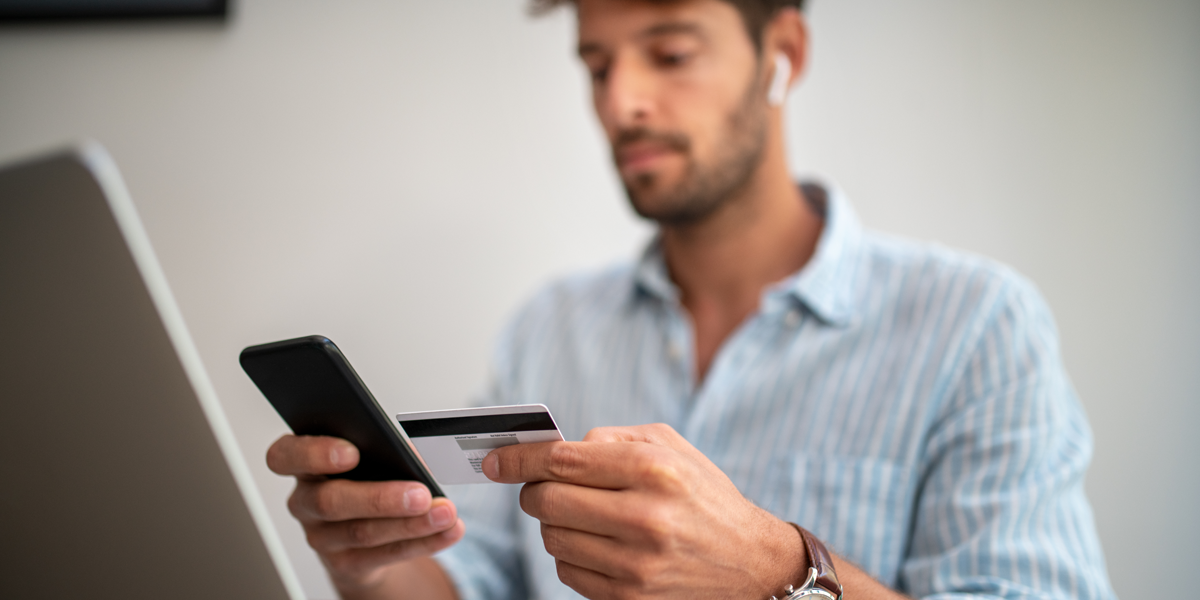 A close-up image of a man holding his phone and credit card while in front of his computer.