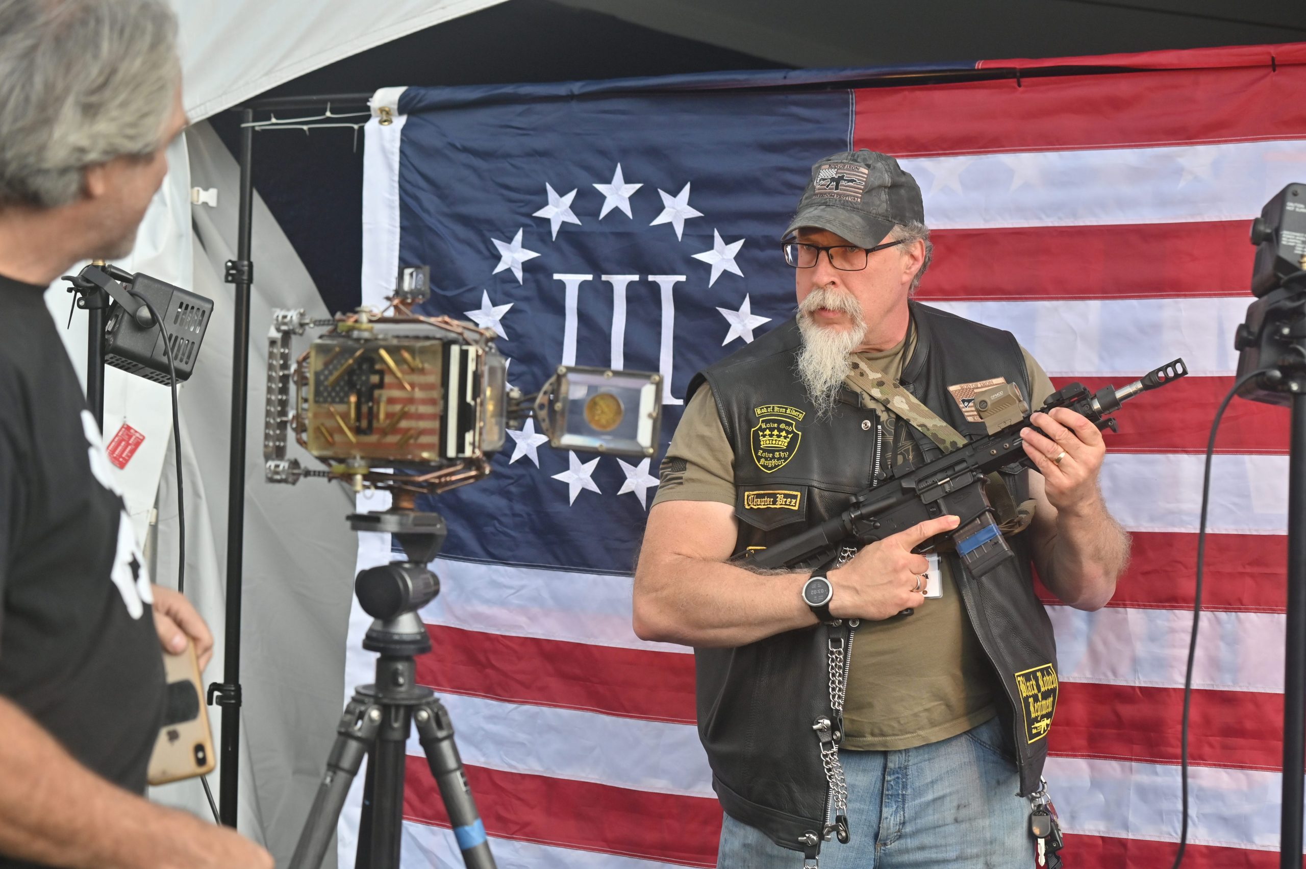 The Rod of Iron Freedom Festival 2021: A man holding an automatic rifle poses in front of a Three Percenter flag, which is a far-right anti-government militia movement.