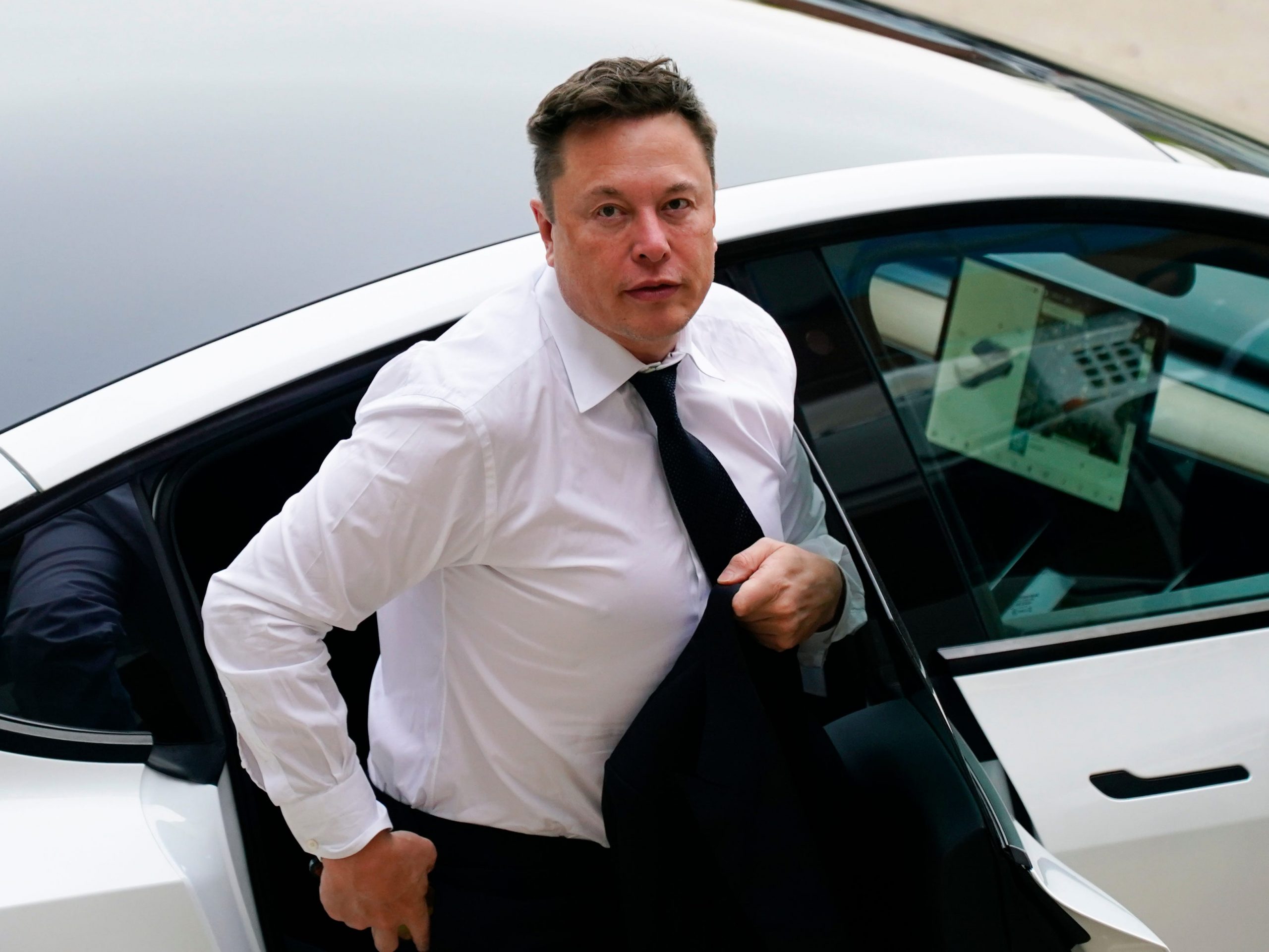 Tesla CEO Elon Musk stepping out of a silver Tesla wearing a white shirt and black tie on a sunny day