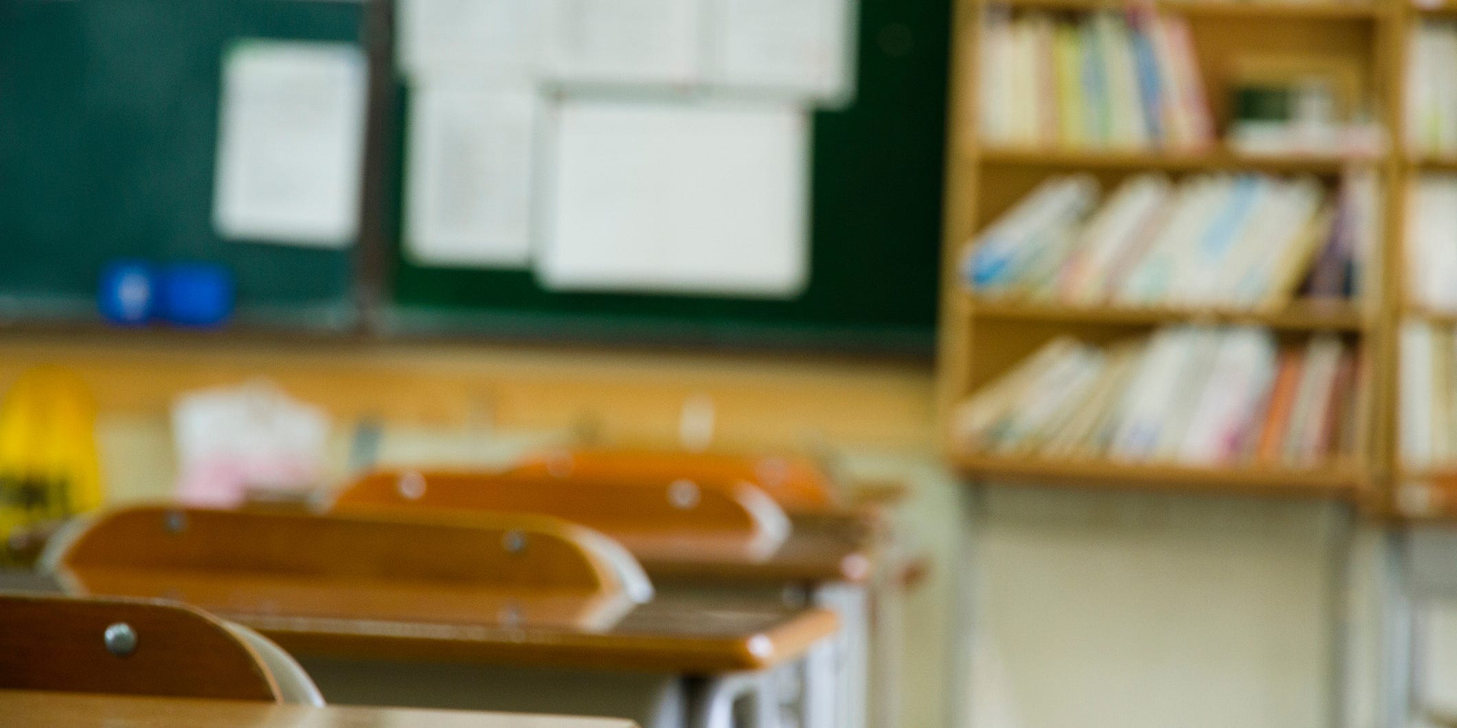 A school classroom, with desks, a blackboard, and bookshelves.