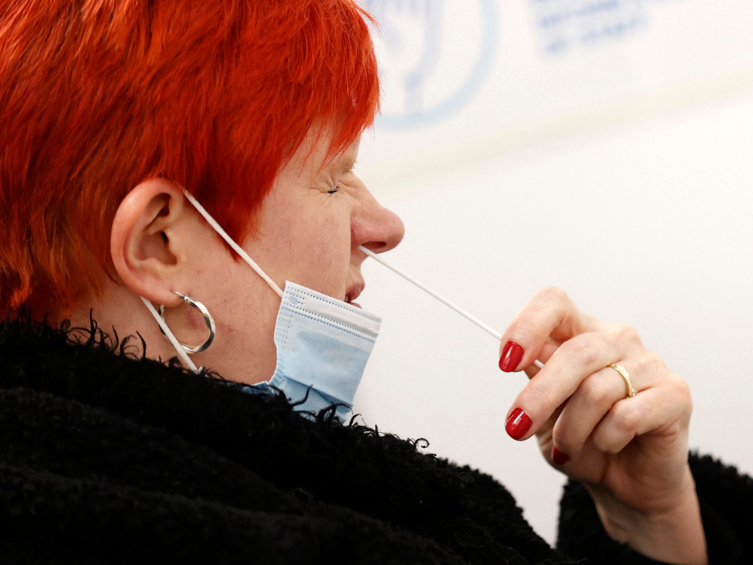 Women with red hair and red nail varnish in a black coat and gold hoops puts COVID-19 test swab into right nostril.
