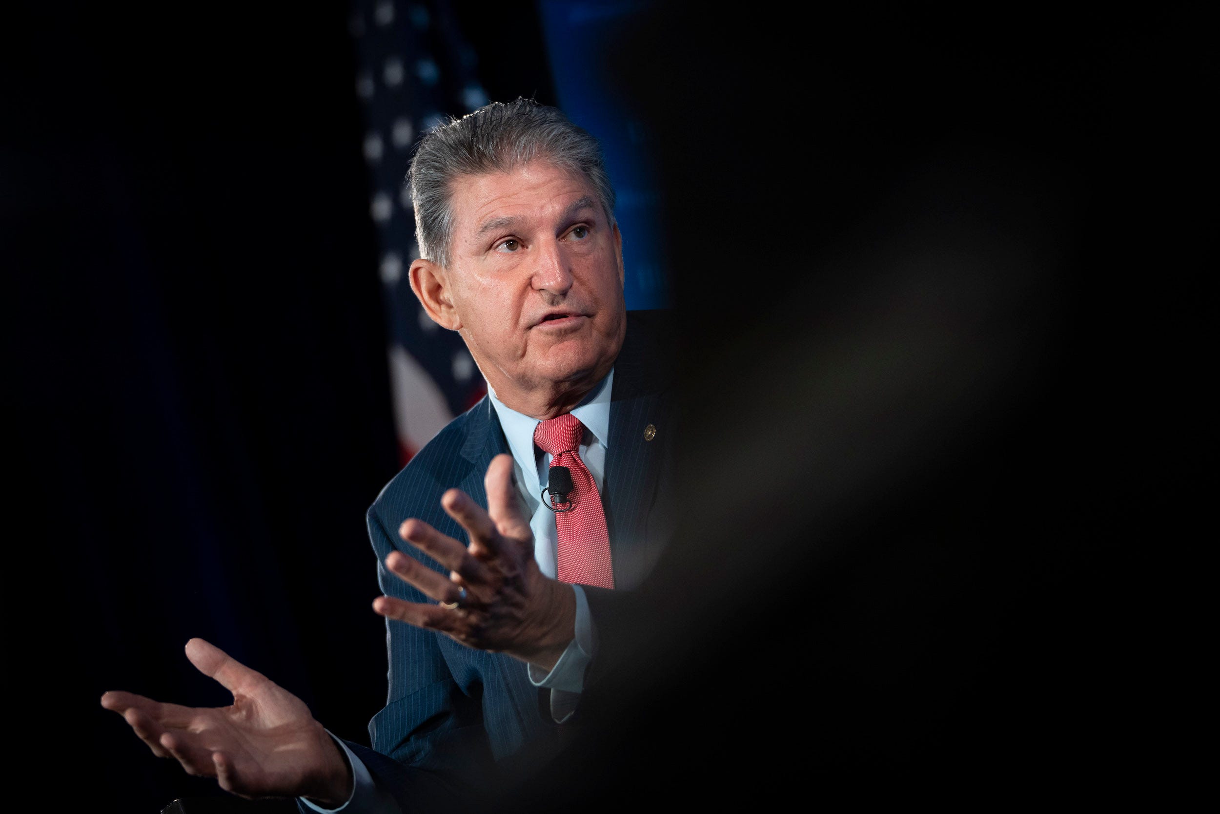 Senator Joe Manchin gestures in front of an American flag with his palms up, the right half of the photo is blurred out in black.