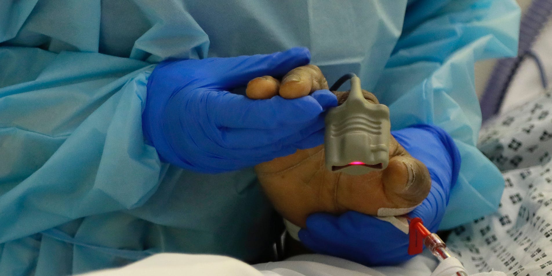 A close-up of a UK medical worker in PPE holding the hand of a COVID-19 patient, who has an oxygen monitor on their finger.