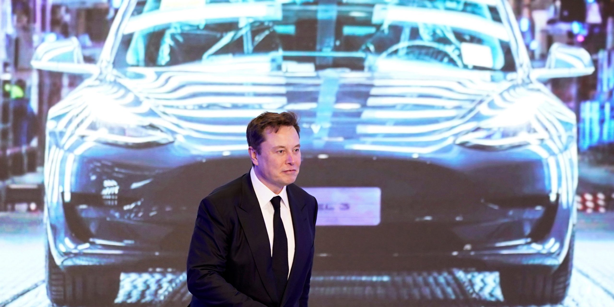 Tesla CEO Elon Musk in a black suit walks on stage in front of an image of a Model Y vehicle