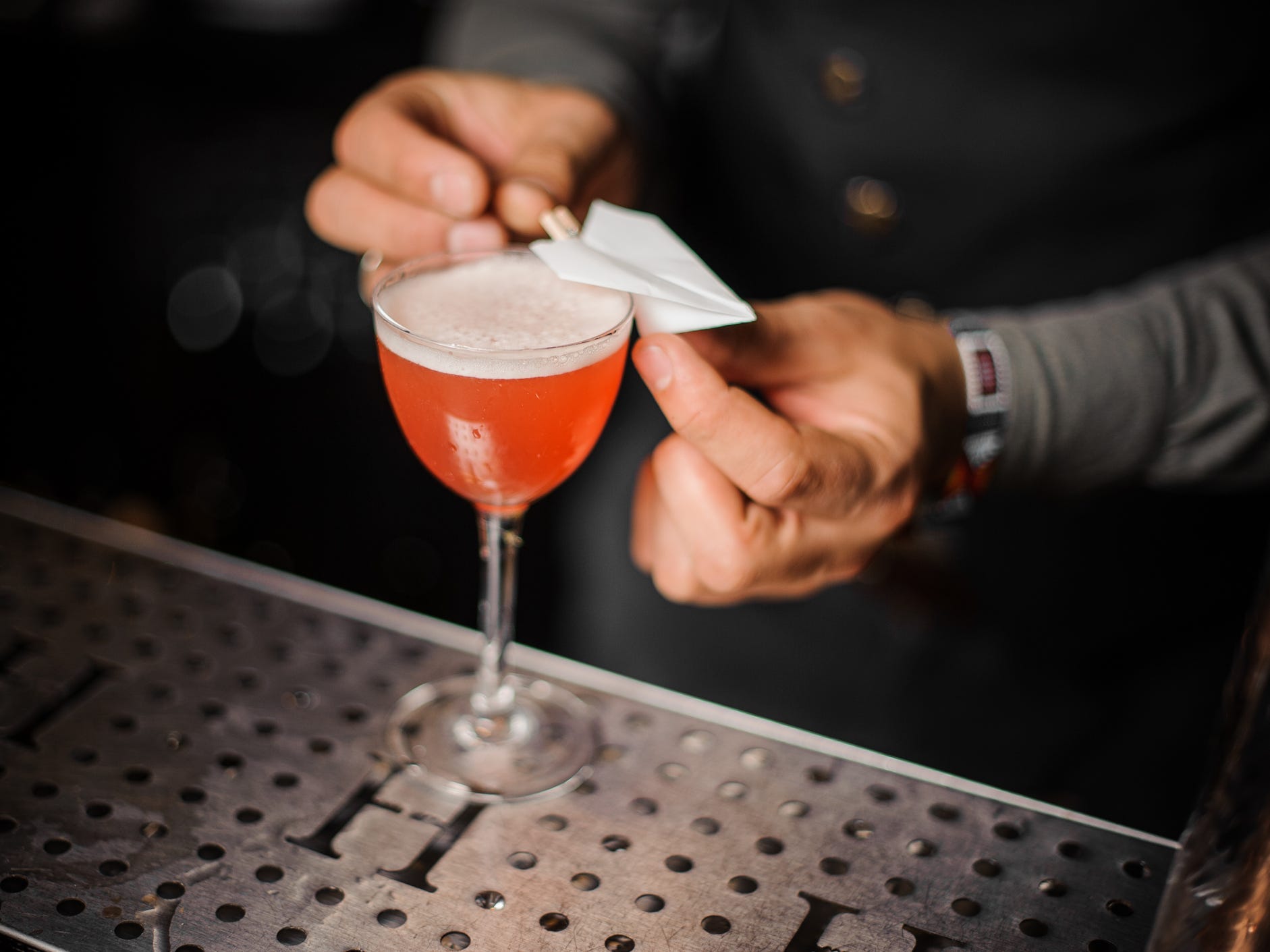 A bartender garnishing a paper plane cocktail with an actual small paper plane