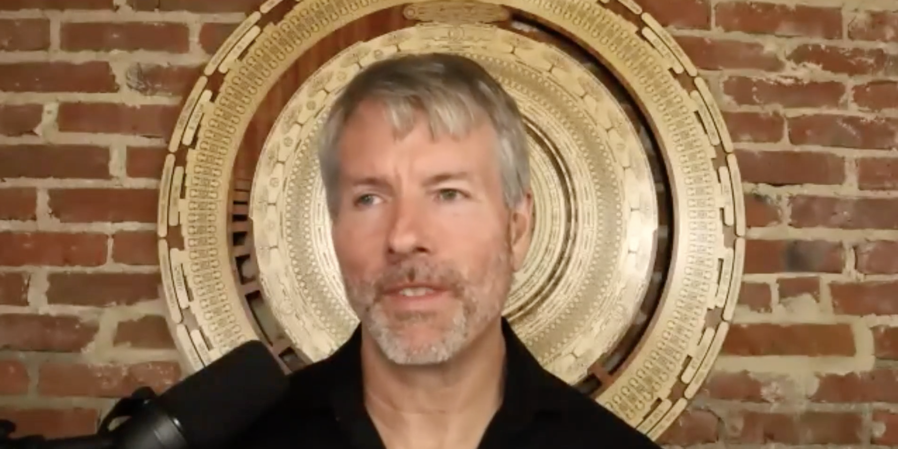 Michael Saylor, speaks into a microphone, wearing a black shirt in front of a brick wall featuring a large gold bitcoin sculpture.