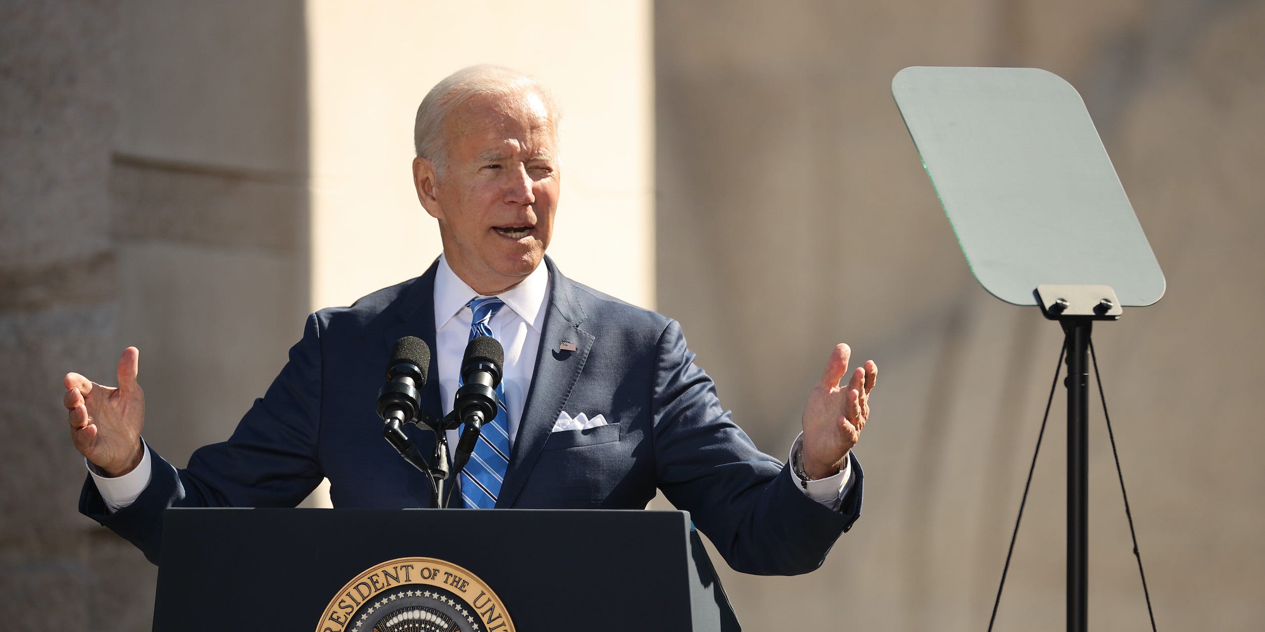 President Joe Biden speaks in front of the Martin Luther King Jr. memorial, gesturing with open palms to his sides while looking toward a teleprompter.