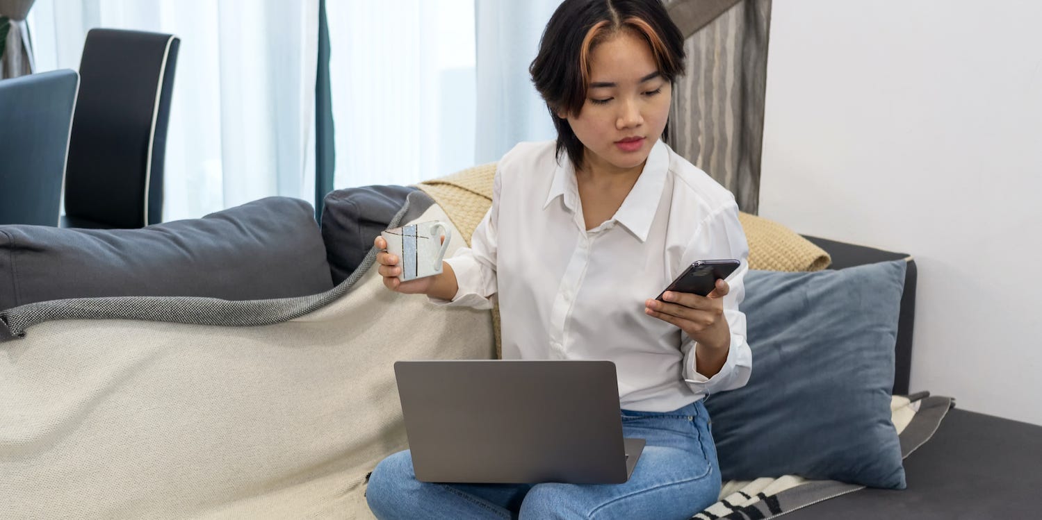 Young woman looking at cell phone with laptop on her lap.