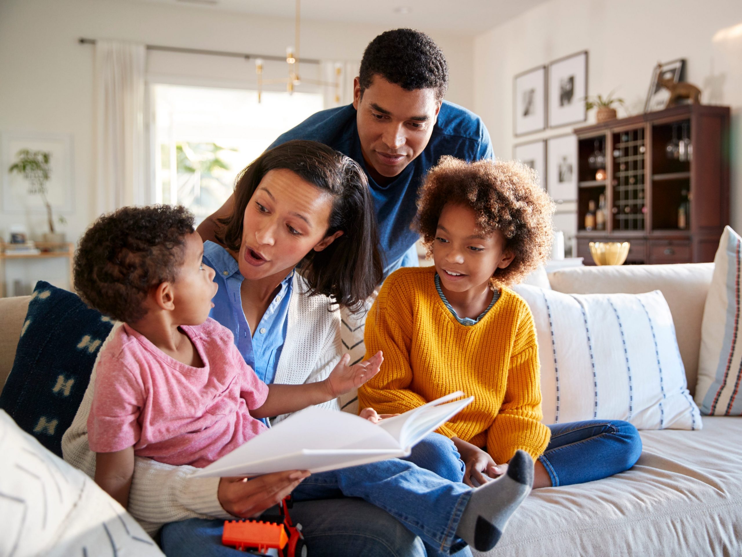 Young mother sitting on sofa in the living room with her daughter beside her and toddler son on her knee, father standing behind them