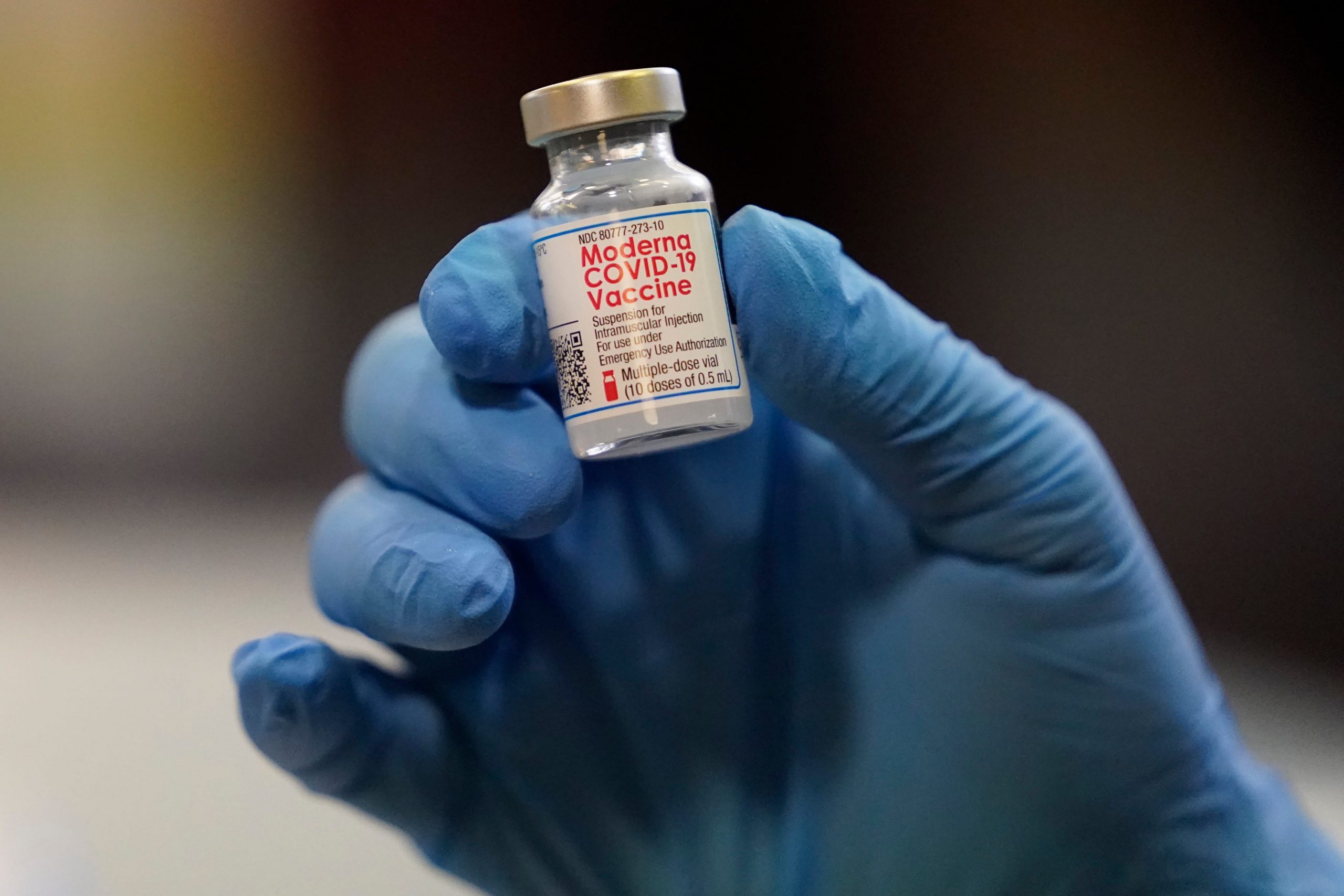 A glass vial of Moderna COVID-19 Vaccine held by a hand in a blue surgical glove