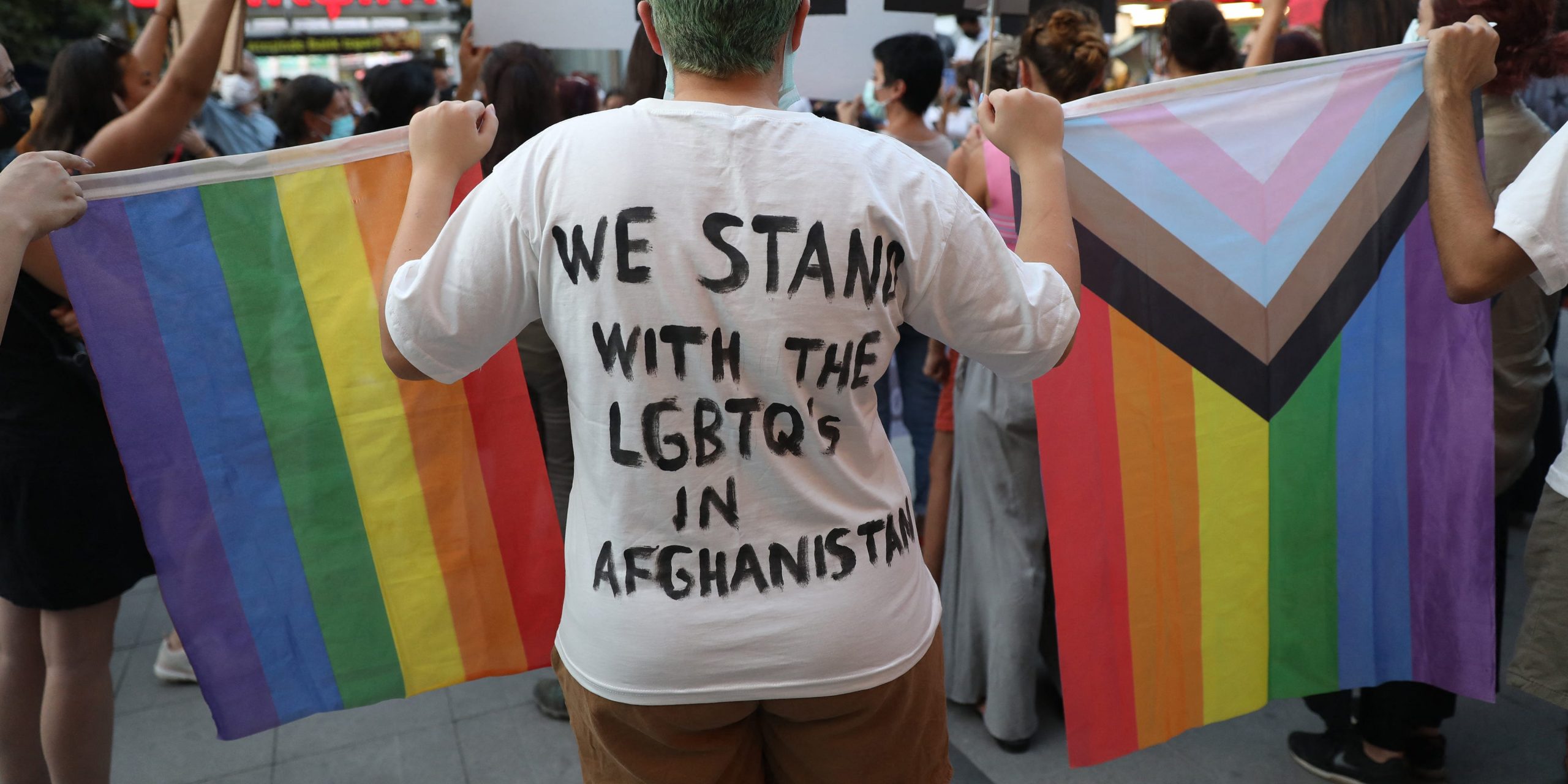 A person holds a rainbow flag and wears a shirt that says, "We stand with the LGBTQs in Afghanistan."