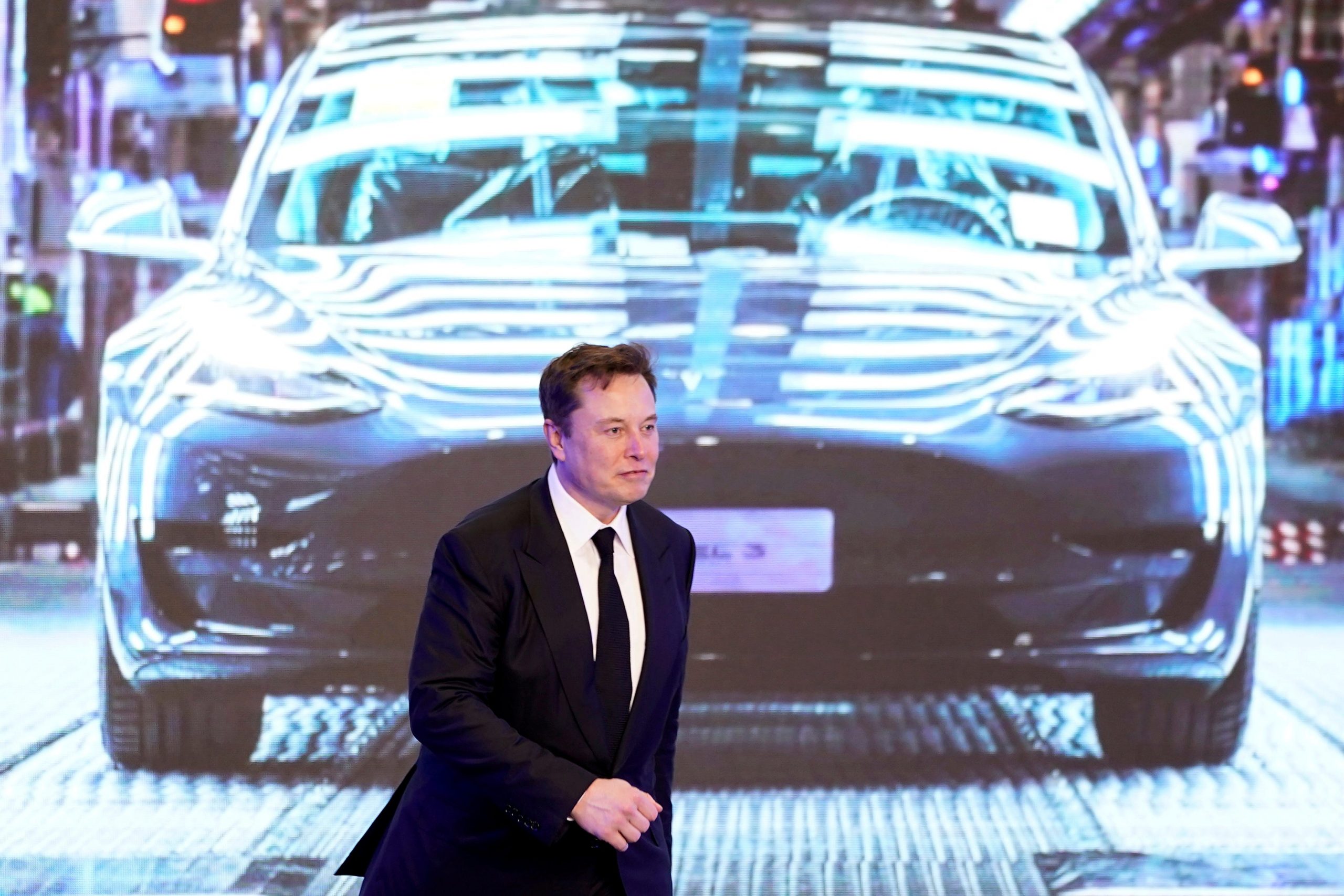 Tesla CEO Elon Musk in a black suit walks on stage in front of an image of a Model Y vehicle