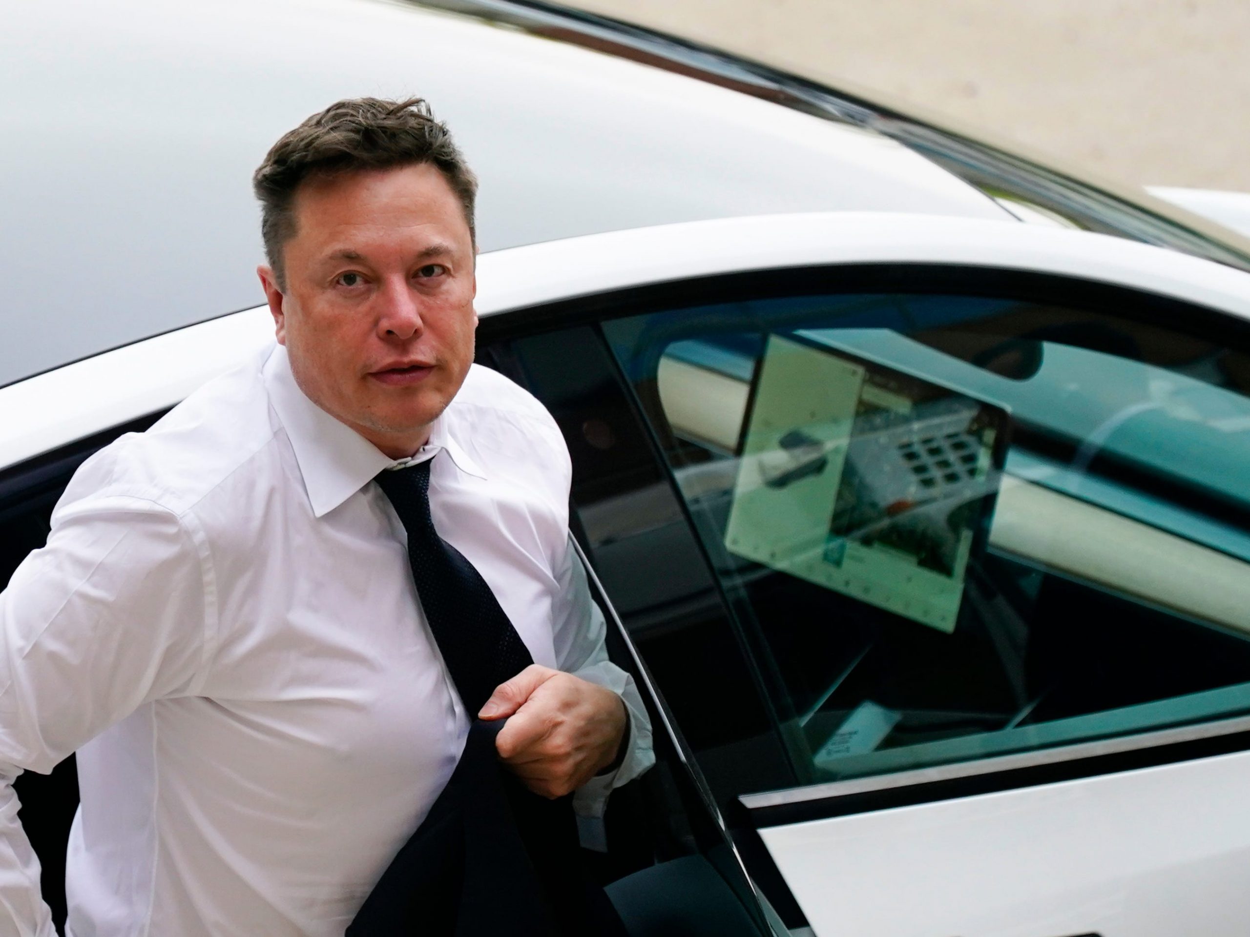 Tesla CEO Elon Musk stepping out of a silver Tesla wearing a white shirt and black tie on a sunny day