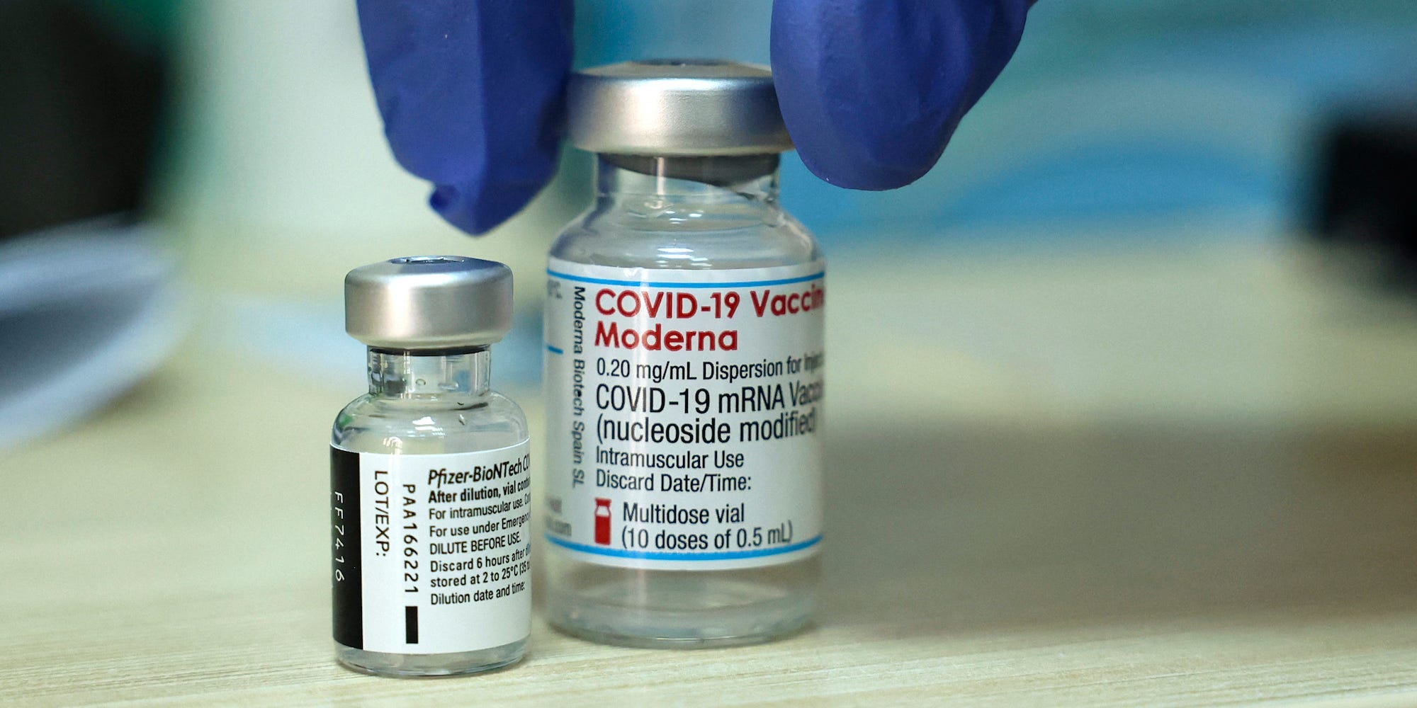 A gloved hand reaches toward two glass vials of Pfizer and Moderna vaccines
